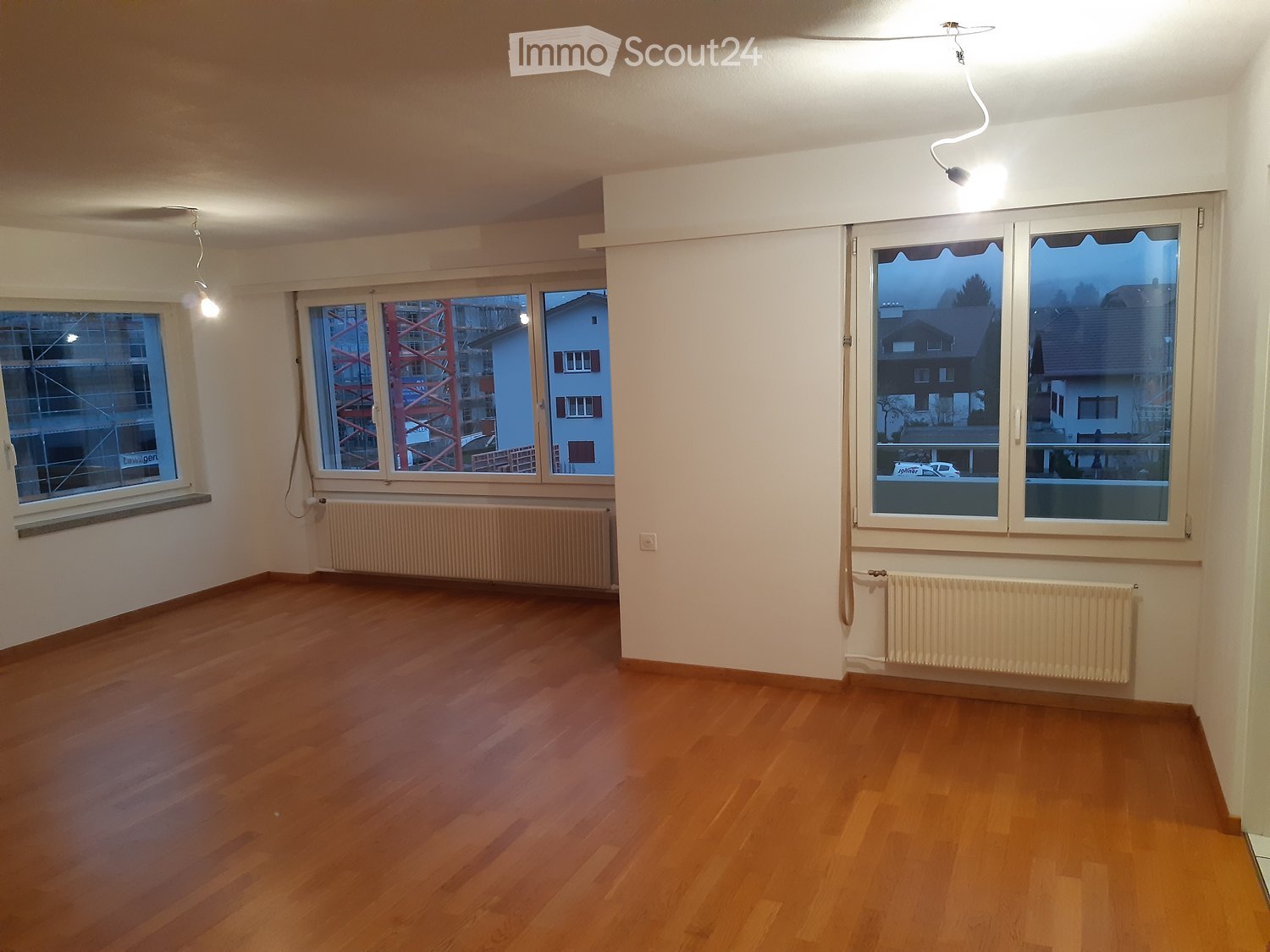 Empty room with wooden flooring, white walls, large windows, radiators, and pendant lights.