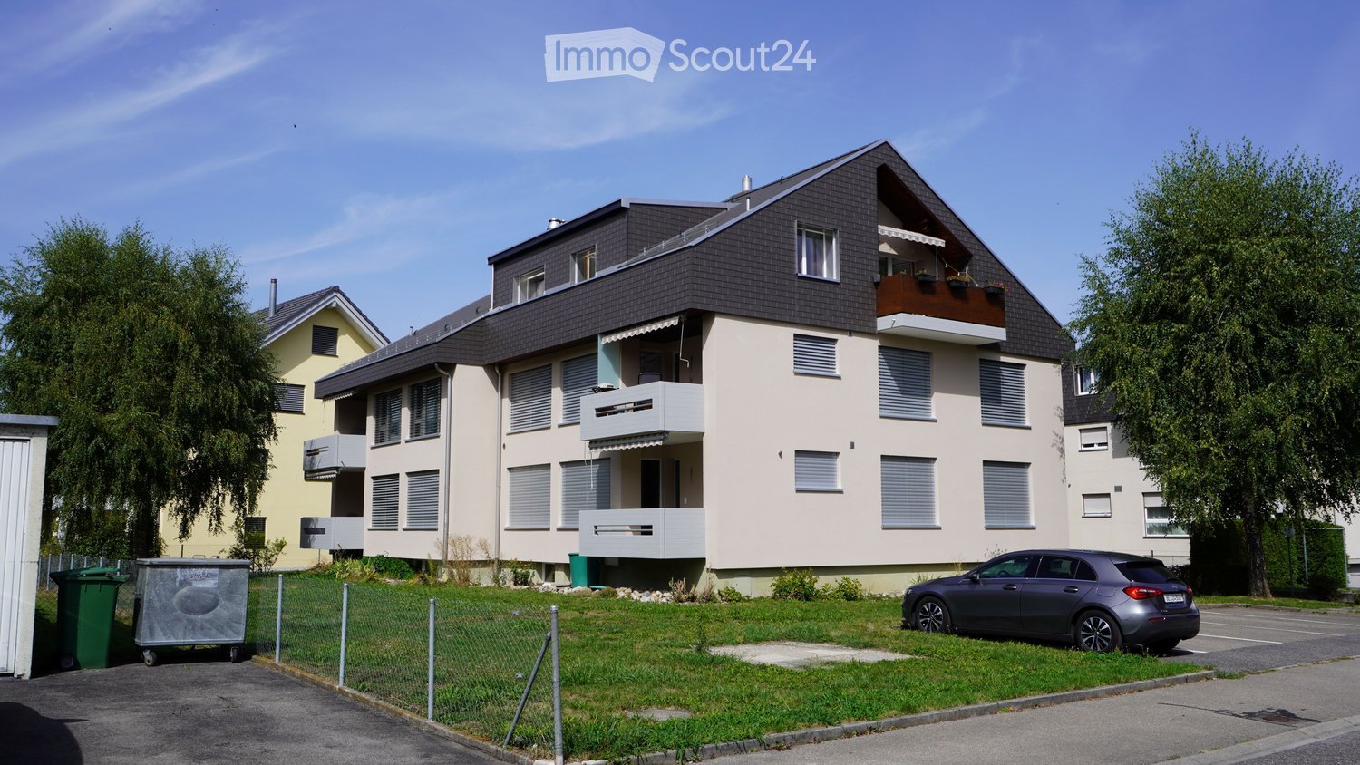 Two-story residential building with beige walls, dark grey roof, multiple windows with shutters, and balconies on the upper floor.