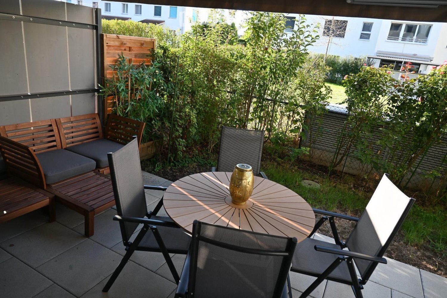 Balcony with wooden bench, round table, and chairs, surrounded by greenery and a decorative vase.