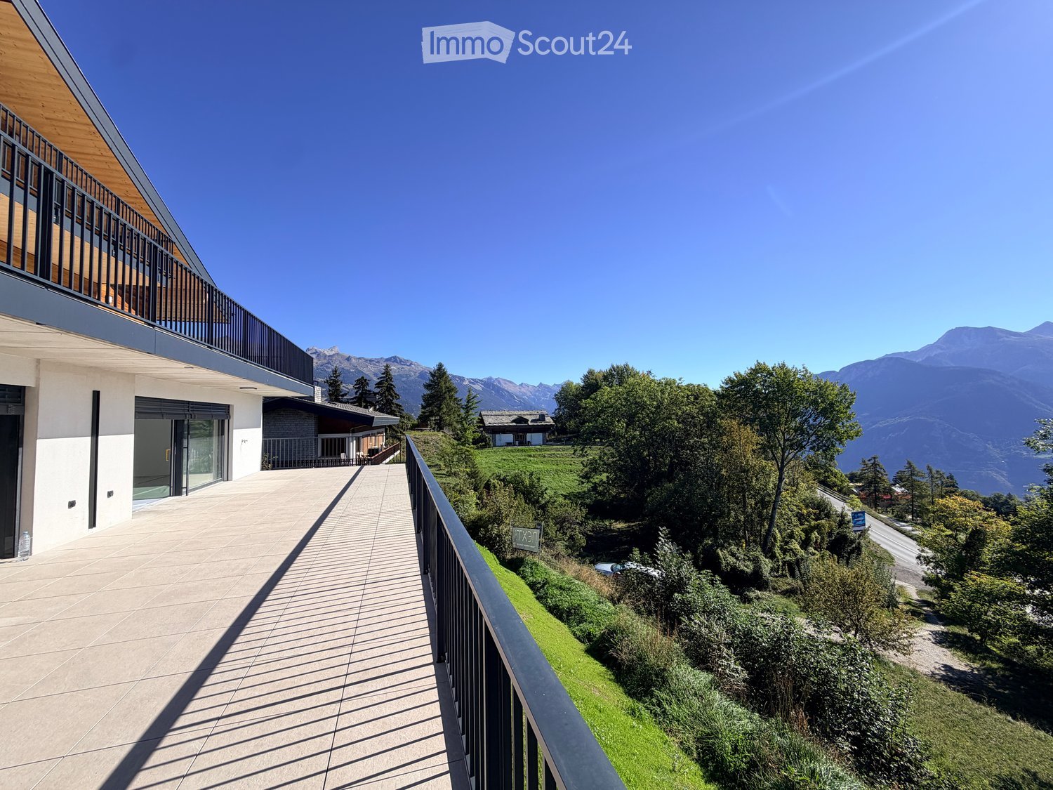 Modern balcony with wooden decking, black metal railing, overlooking a lush green hillside and distant mountains.