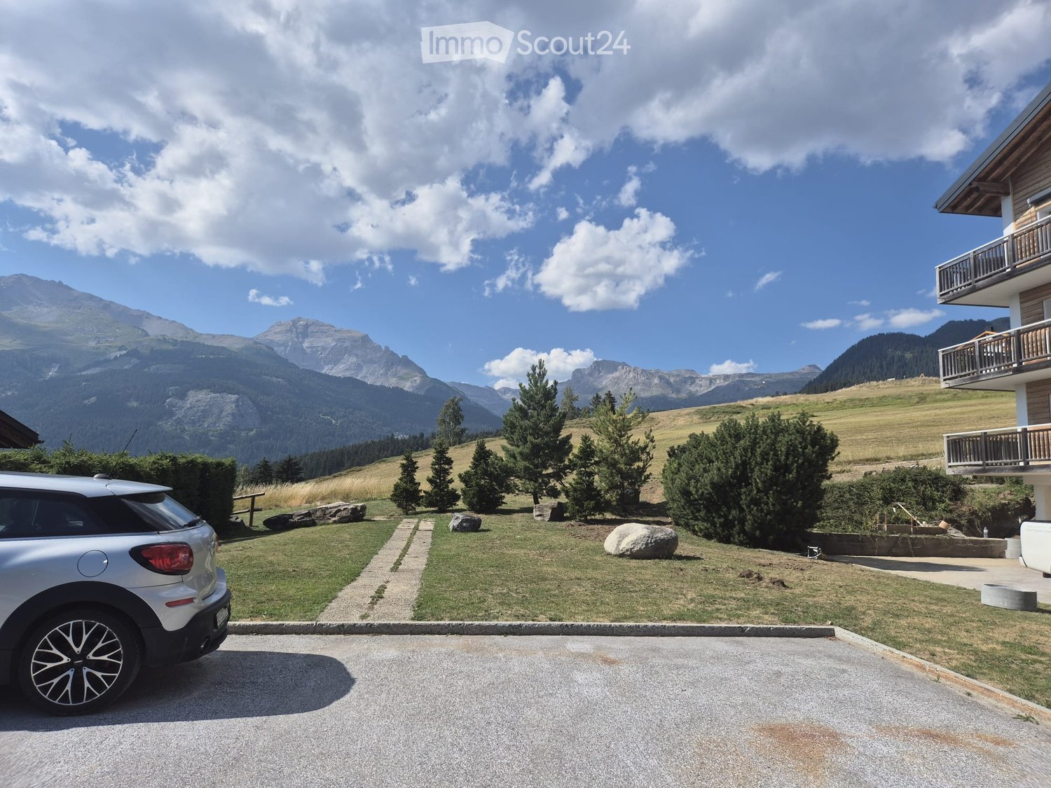 A parking area with a silver car, surrounded by a grassy lawn, trees, and a large building with a balcony. The background features a scenic mountain range under a partly cloudy sky.