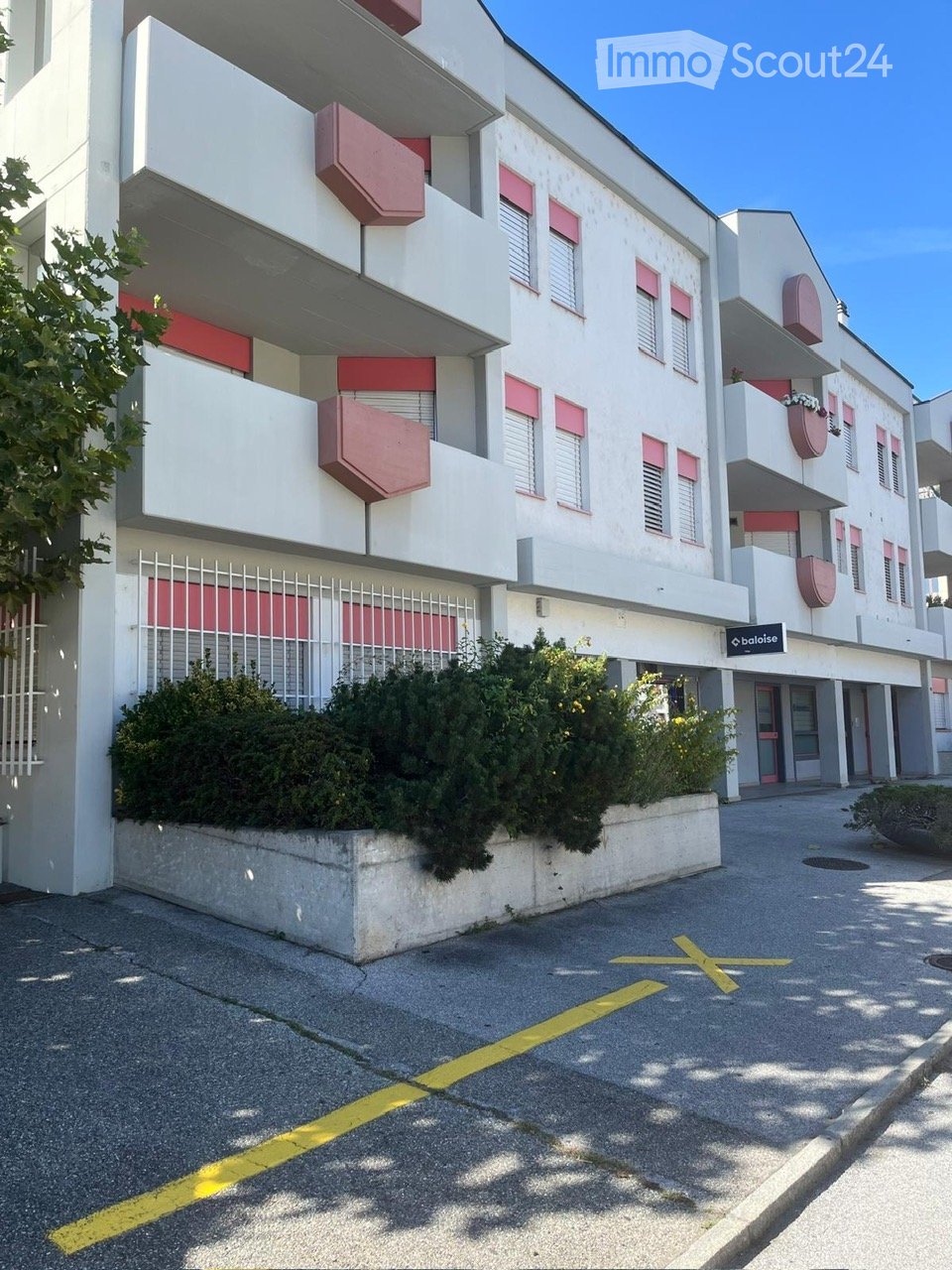 Modern multi-story building with white and red accents, balconies, and greenery in front.