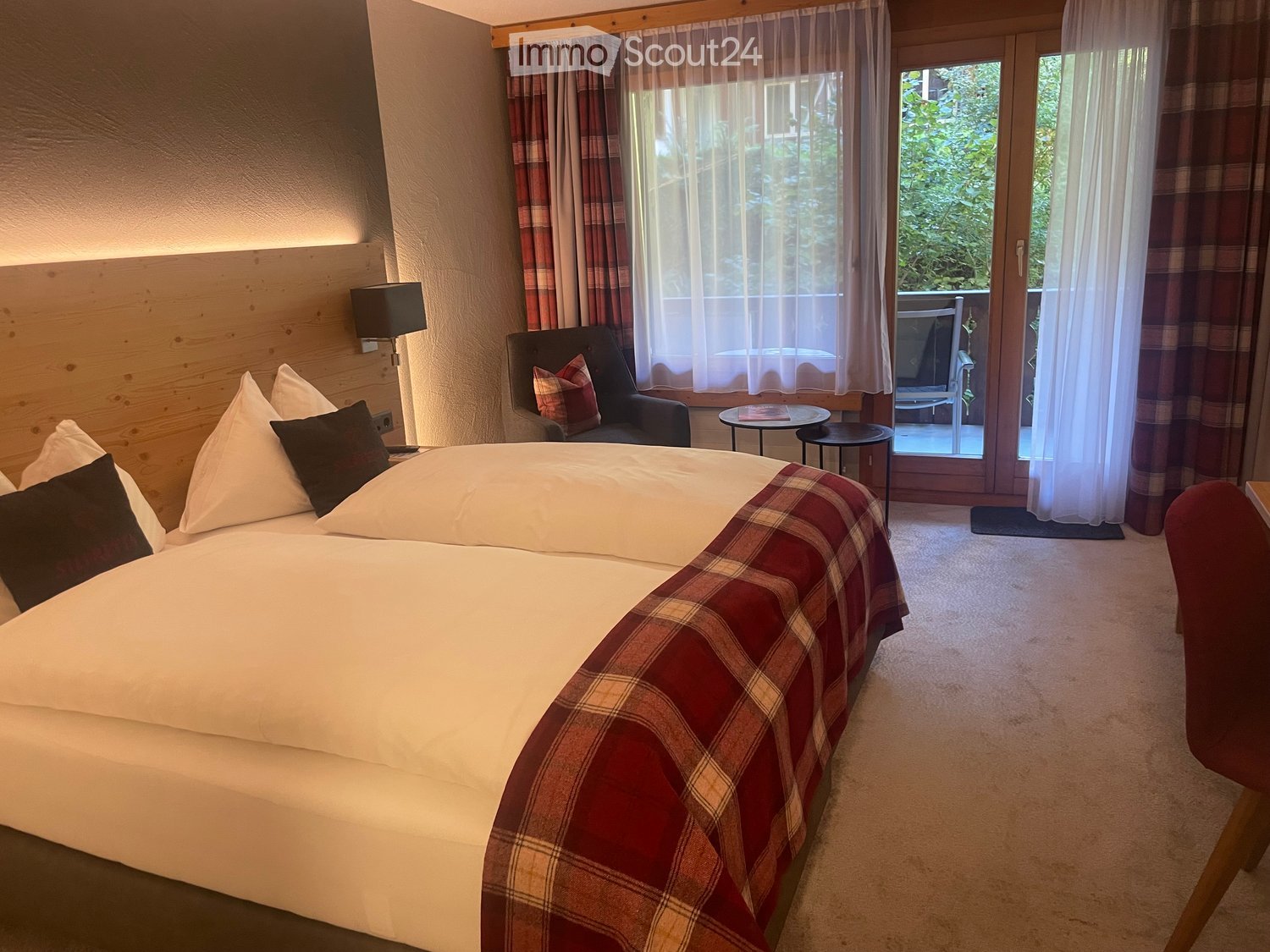 Cozy bedroom with a large bed, plaid blanket, armchair, small round table, and large windows with curtains leading to a balcony.