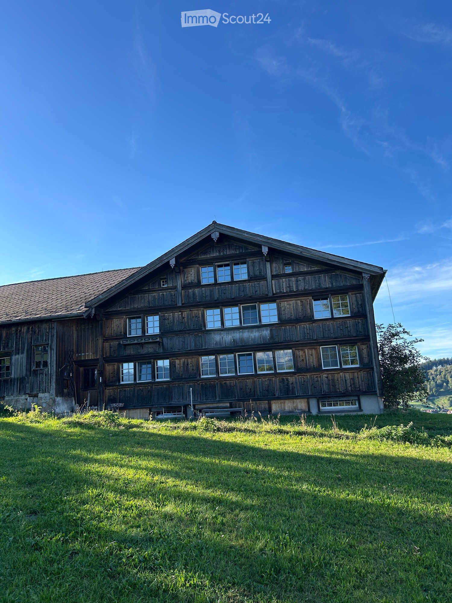 A two-story wooden house with a steeply pitched roof, multiple windows, and a stone foundation, situated on a grassy lawn with a scenic mountain backdrop.