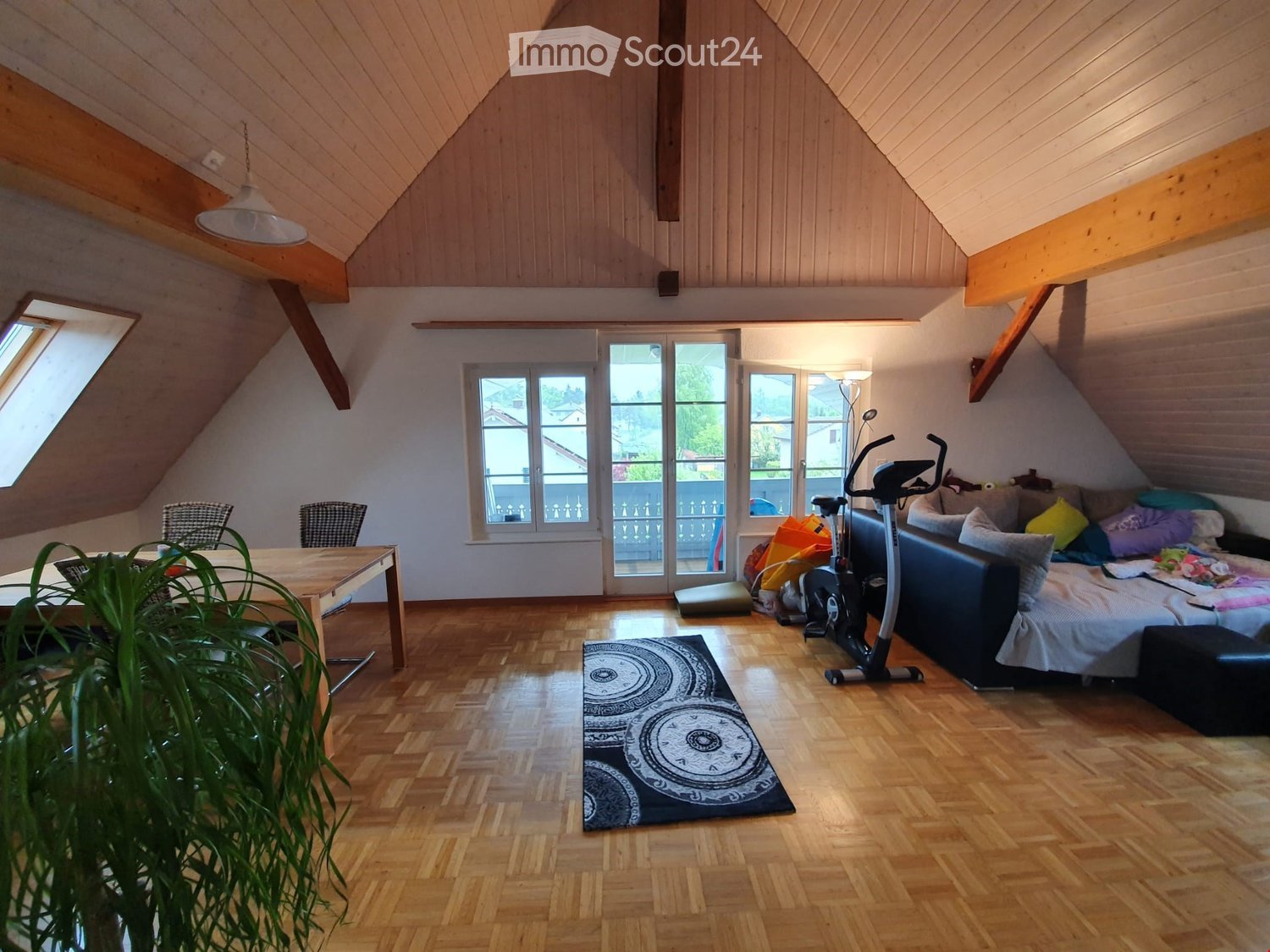 Bright attic living space with wooden floors, slanted wooden ceiling, large windows, and a sliding glass door leading to a balcony. Features a sofa, exercise bike, dining table, chairs, and a patterned rug.