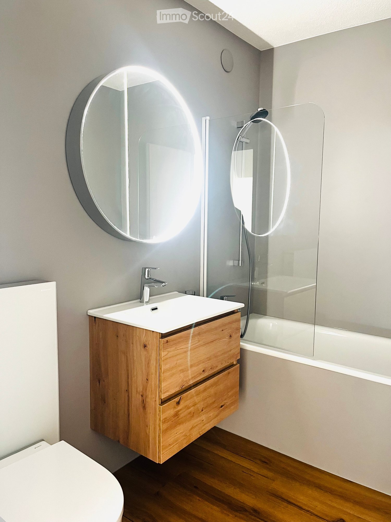 Modern bathroom with a wooden vanity, white sink, chrome faucet, large round mirror, and a glass-enclosed bathtub.