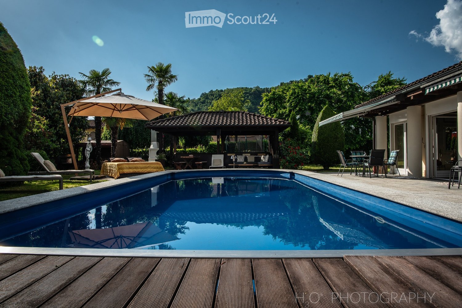 Swimming pool with wooden decking, outdoor seating with umbrella, outdoor dining table, gazebo, lawn with lounge chairs, and tropical trees.