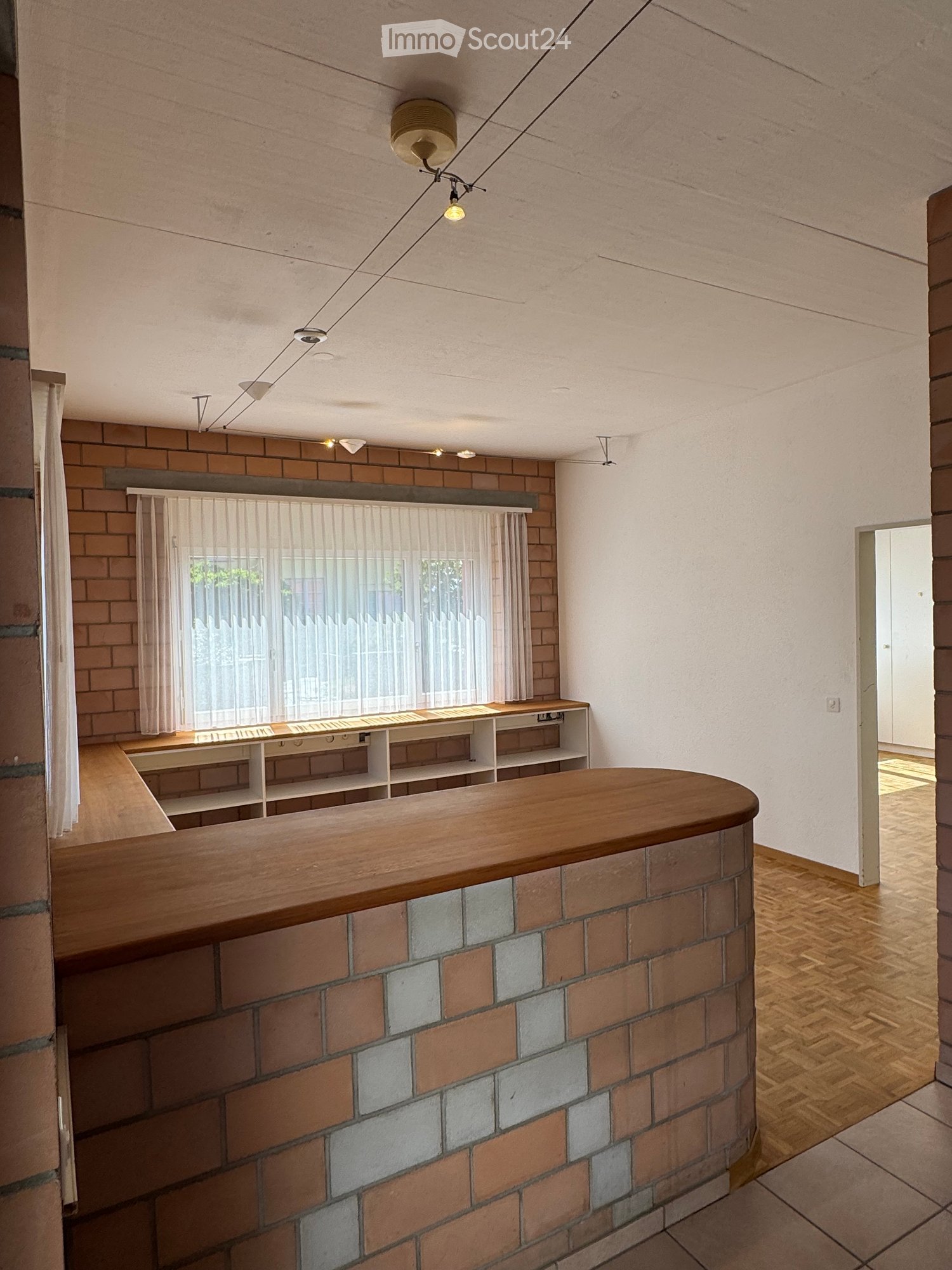 An unfurnished room with exposed brick walls, wooden flooring, and a large window with sheer curtains. The room features built-in shelving units and a curved wooden counter.