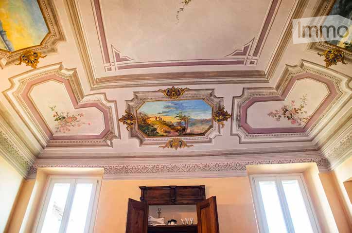 Elegant room with ornate ceiling featuring frescoes and decorative moldings, large windows, and a wooden cabinet with a small table and chair inside.