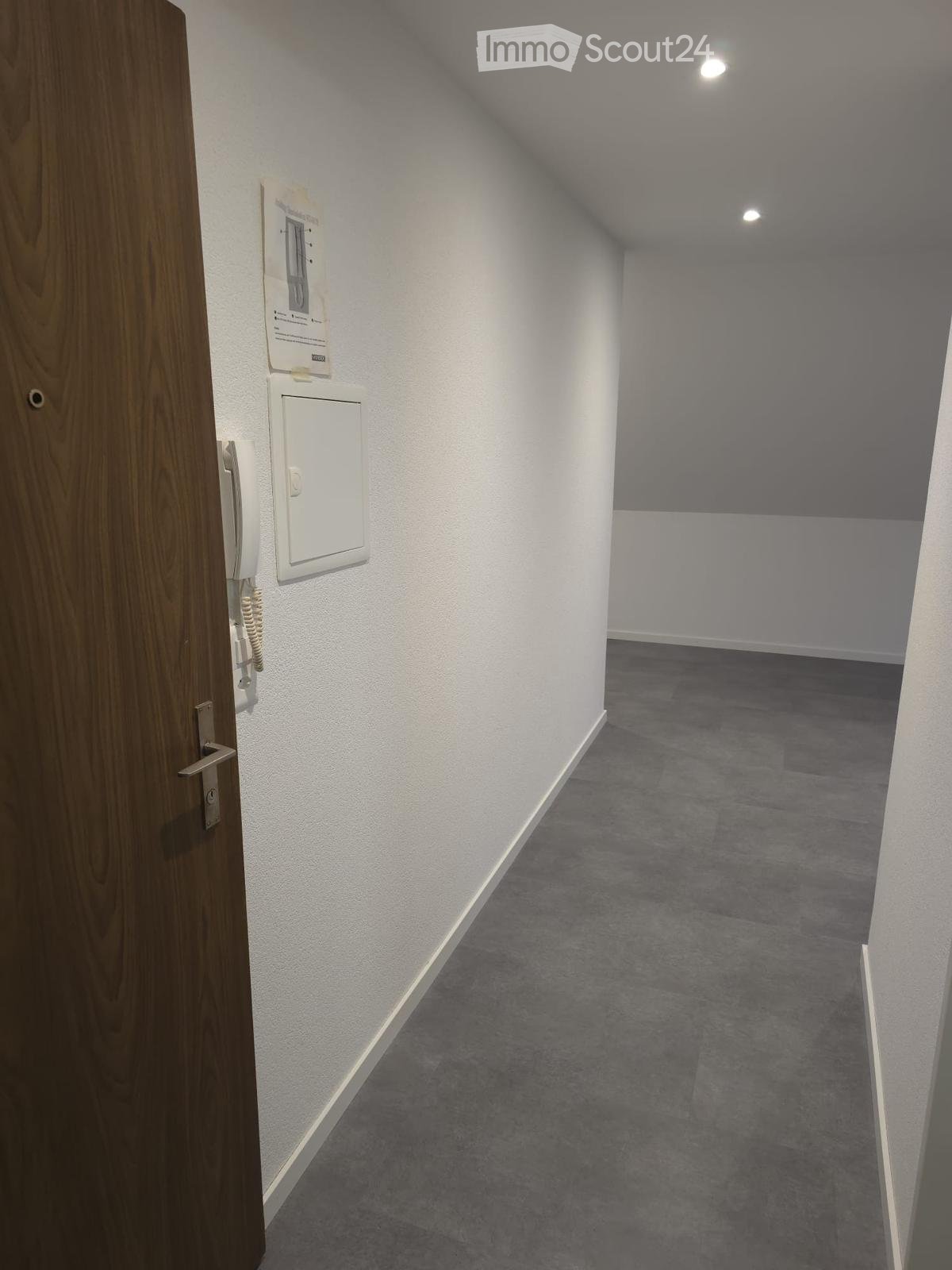 Empty hallway with grey carpet, white walls, and a wooden door on the left. A small intercom system is mounted on the wall near the door.