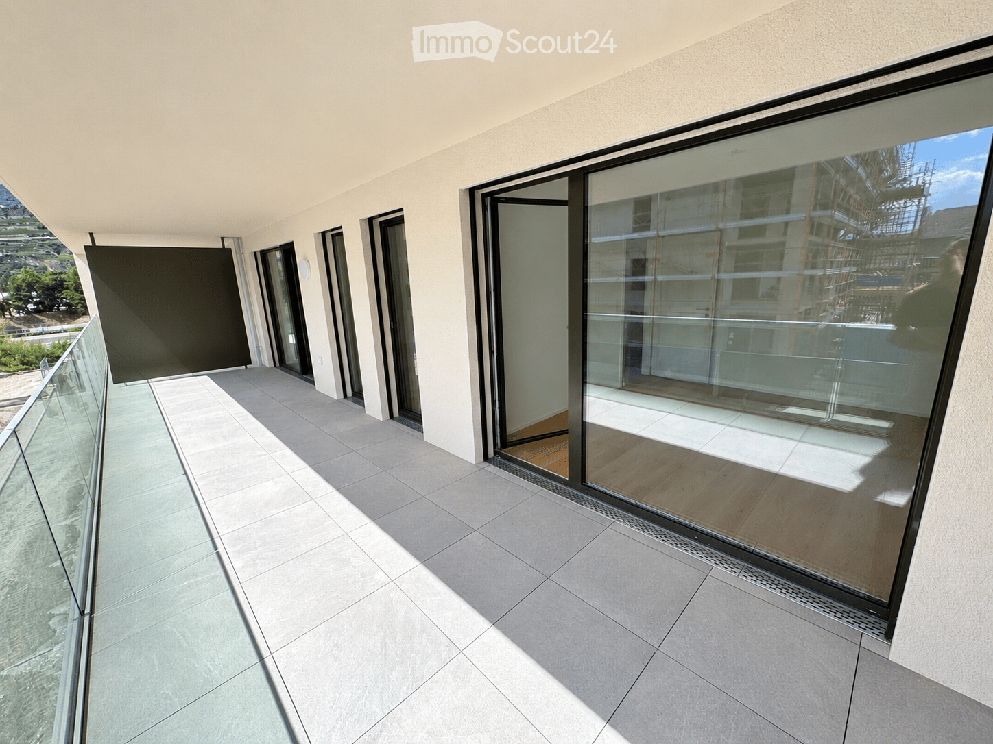 Modern balcony with large glass sliding doors, tiled floor, glass railing, and a view of the surrounding area.