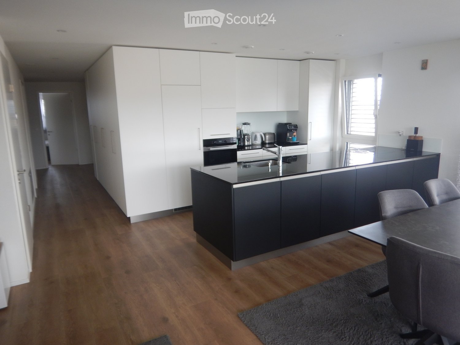 Modern kitchen with white and dark grey cabinets, a central island, and a dining area with a table and chairs. The floor is wooden, and the kitchen features a sink, oven, coffee maker, and blender.