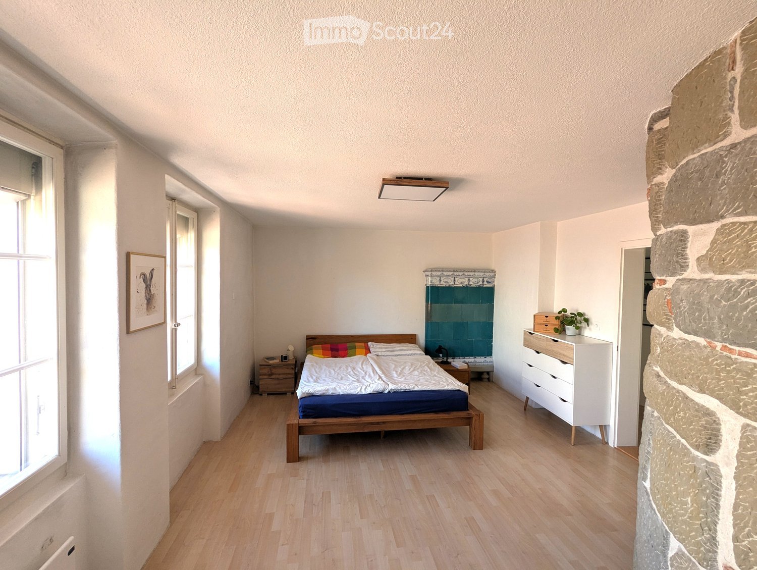 Bright bedroom with wooden flooring, a double bed with colorful bedding, a nightstand with a lamp, a white drawer unit, and a small plant on top.