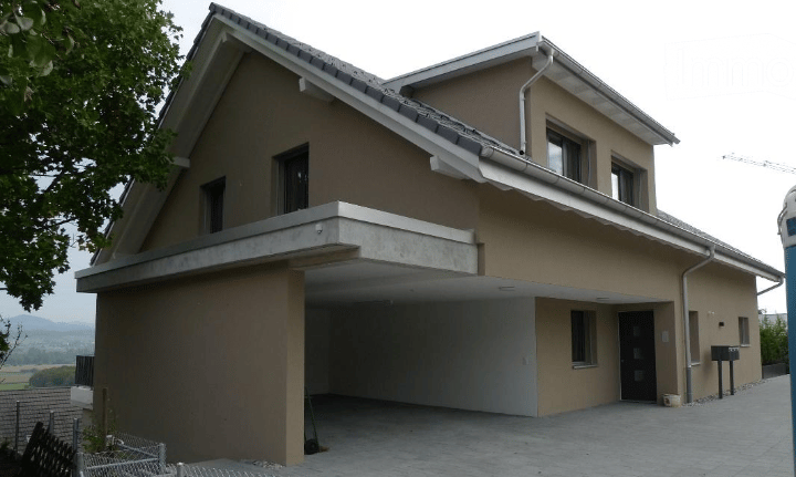 Two-story house with beige exterior, garage, and multiple windows. The house features a modern design with a sloped roof and a raised ground floor.
