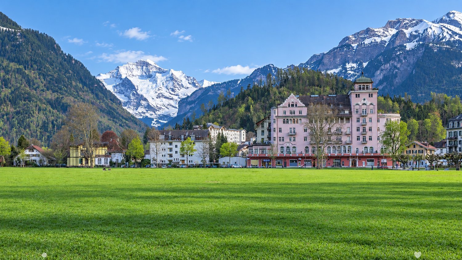 A picturesque town square with a large green lawn, surrounded by colorful buildings and snow-capped mountains in the background.