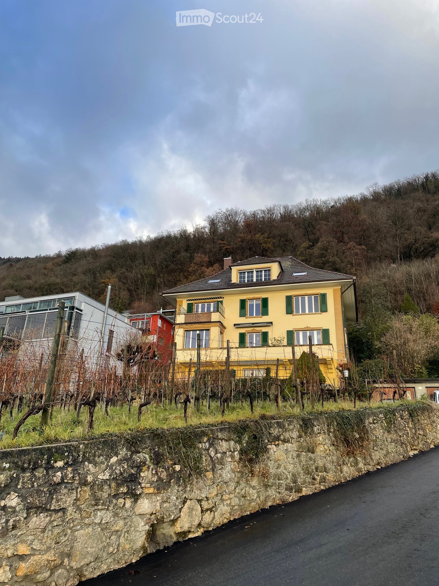 A two-story yellow house with green shutters and a balcony, situated on a hillside with a stone retaining wall and vineyard in the foreground.