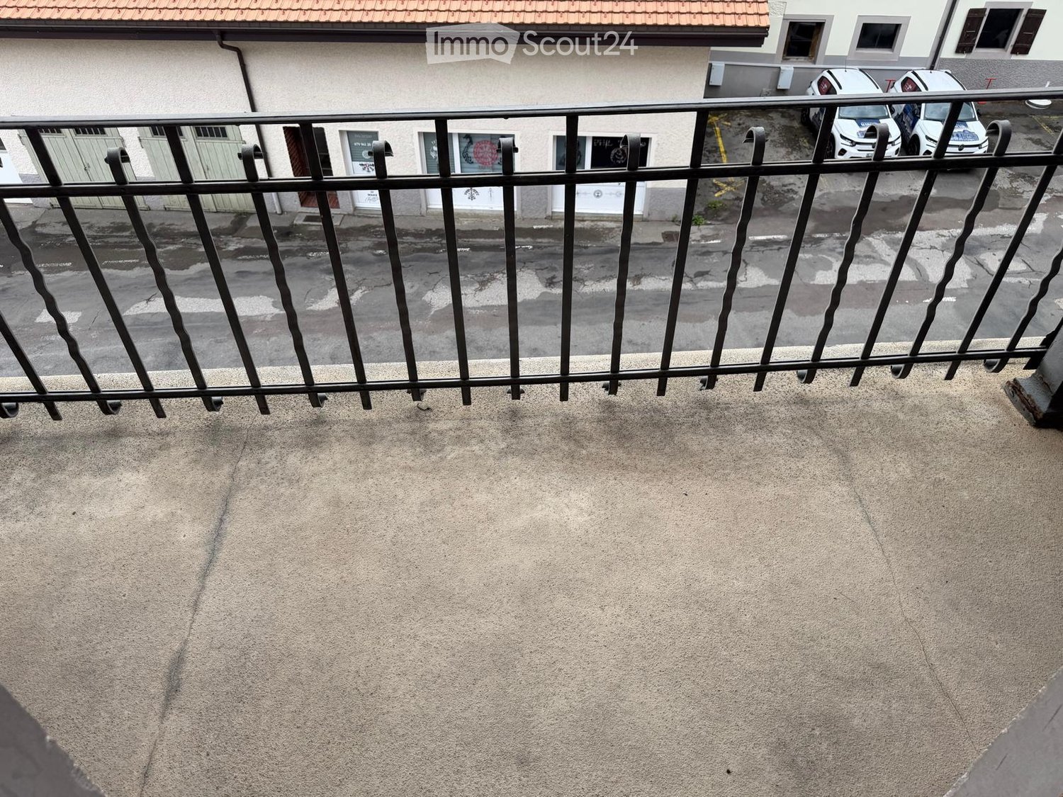 Balcony with metal railing, concrete floor, and view of street with parked cars and buildings.