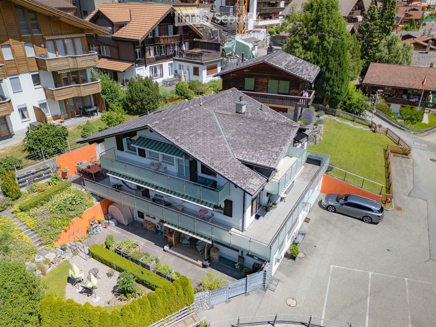 Aerial view of a multi-story house with a grey roof, white walls, and a balcony. The property includes a driveway with a parked car, a well-maintained garden, and outdoor seating areas.