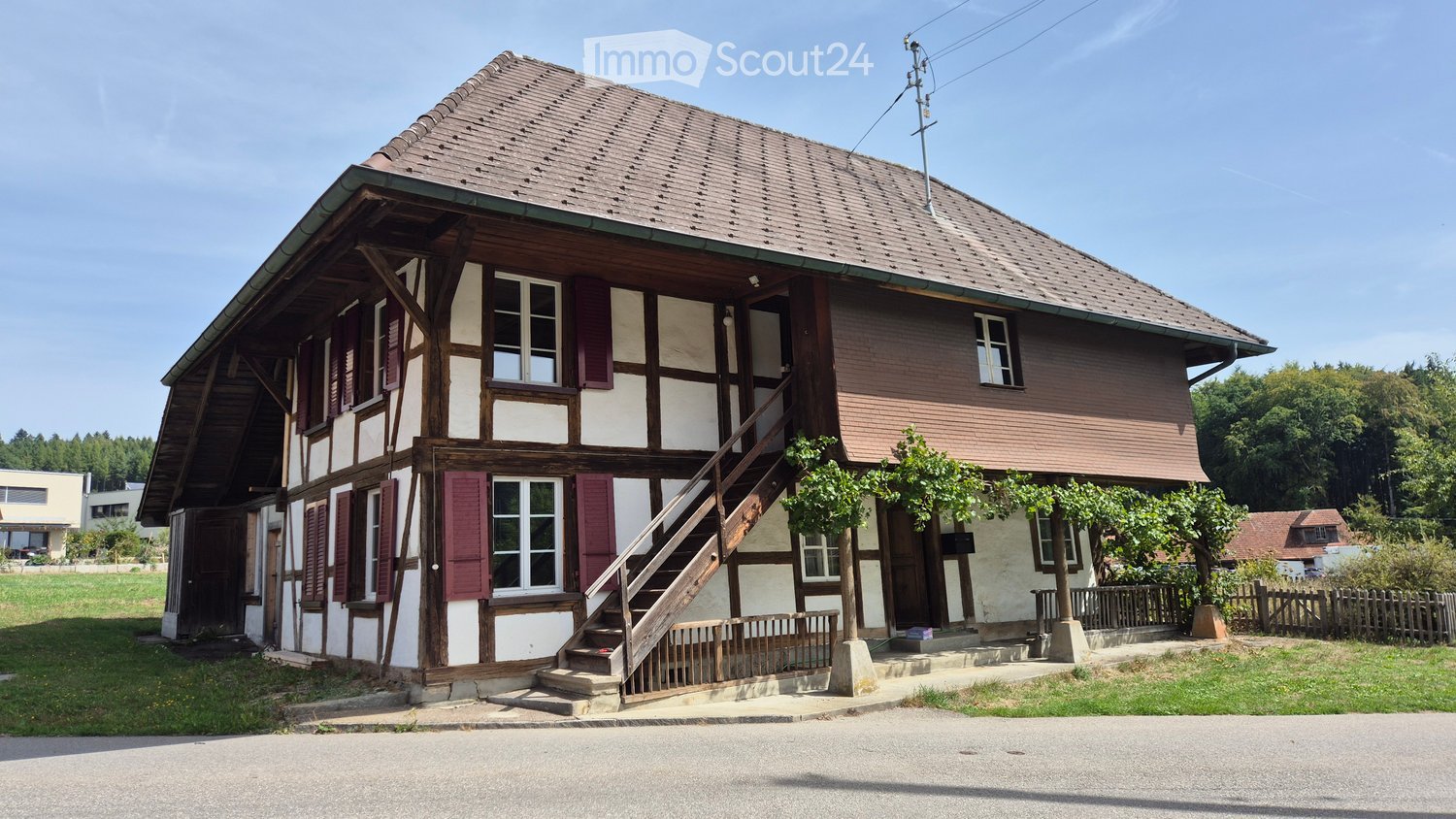 Traditional two-story house with wooden beams, red shutters, and a tiled roof, featuring a wooden staircase leading to the entrance.