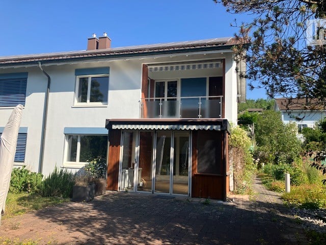 Two-story house with white exterior, blue shutters, and a balcony on the upper floor. The entrance features a covered porch with glass doors.