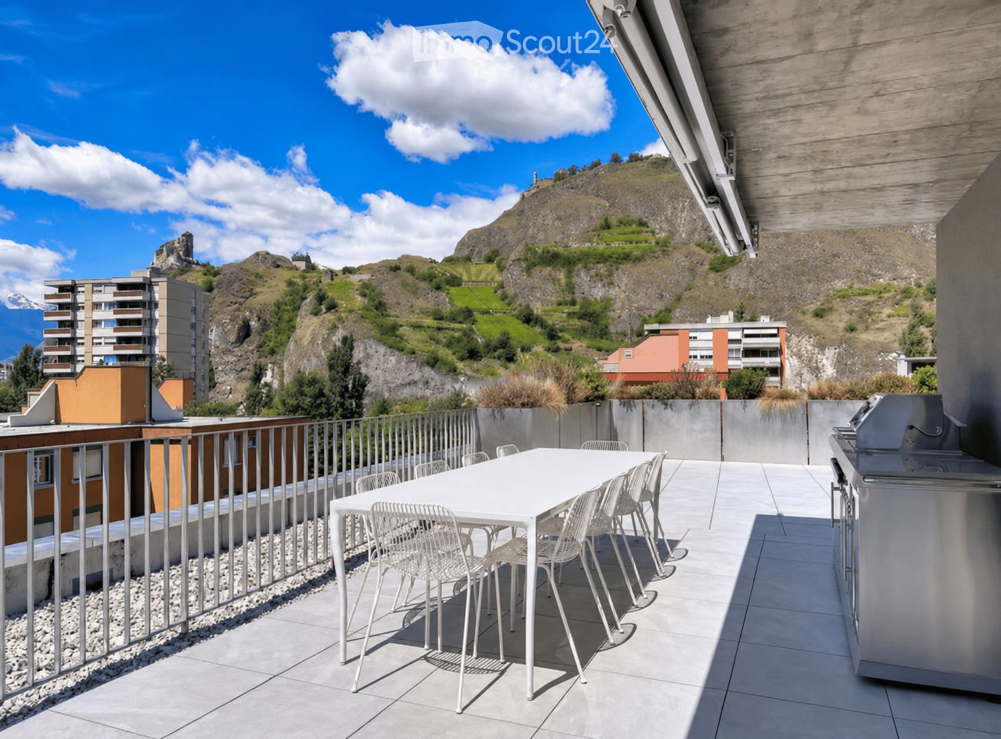 Modern rooftop terrace with a white dining table and chairs, a grill, and a scenic mountain view.