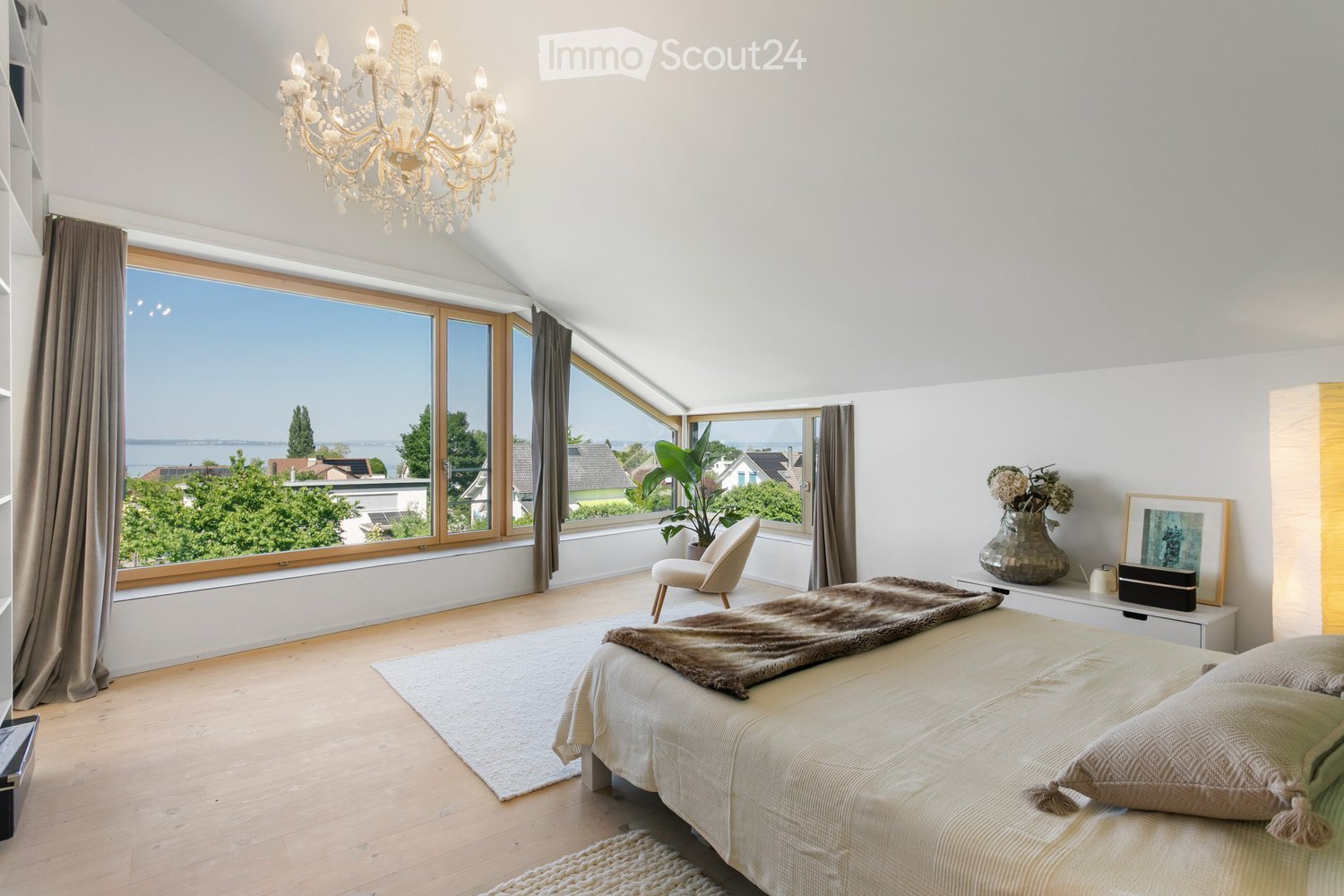 Bright bedroom with large windows offering a scenic view, featuring a bed with a fur throw, a white armchair, a chandelier, and decorative items on a side table.