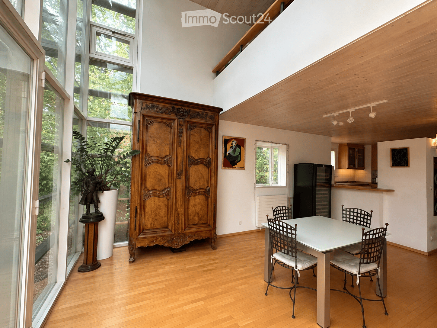 Bright, open room with wooden floors, large windows, a dining table with four chairs, a tall wooden cabinet, a refrigerator, a potted plant, and a decorative statue.