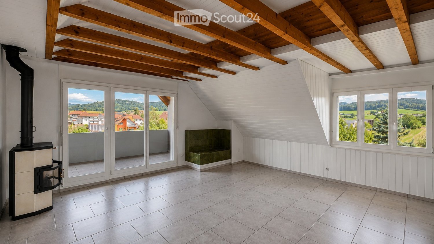 Bright, empty attic room with wooden beams, large windows, and a wood-burning stove. The space features white paneled walls, tiled flooring, and a small seating area with a green cushioned bench.