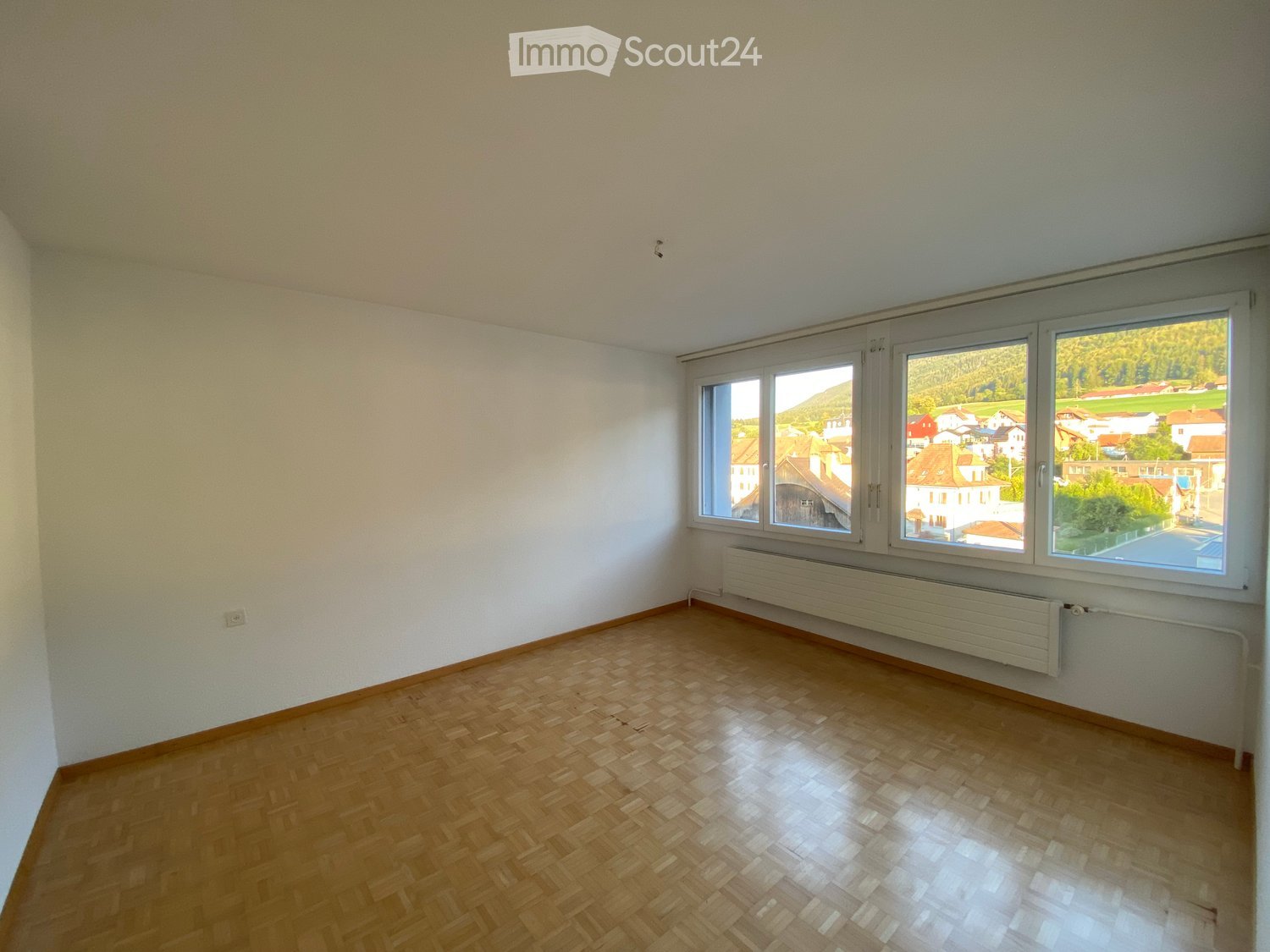 Empty room with wooden flooring, white walls, and large windows offering a view of a town and surrounding landscape.