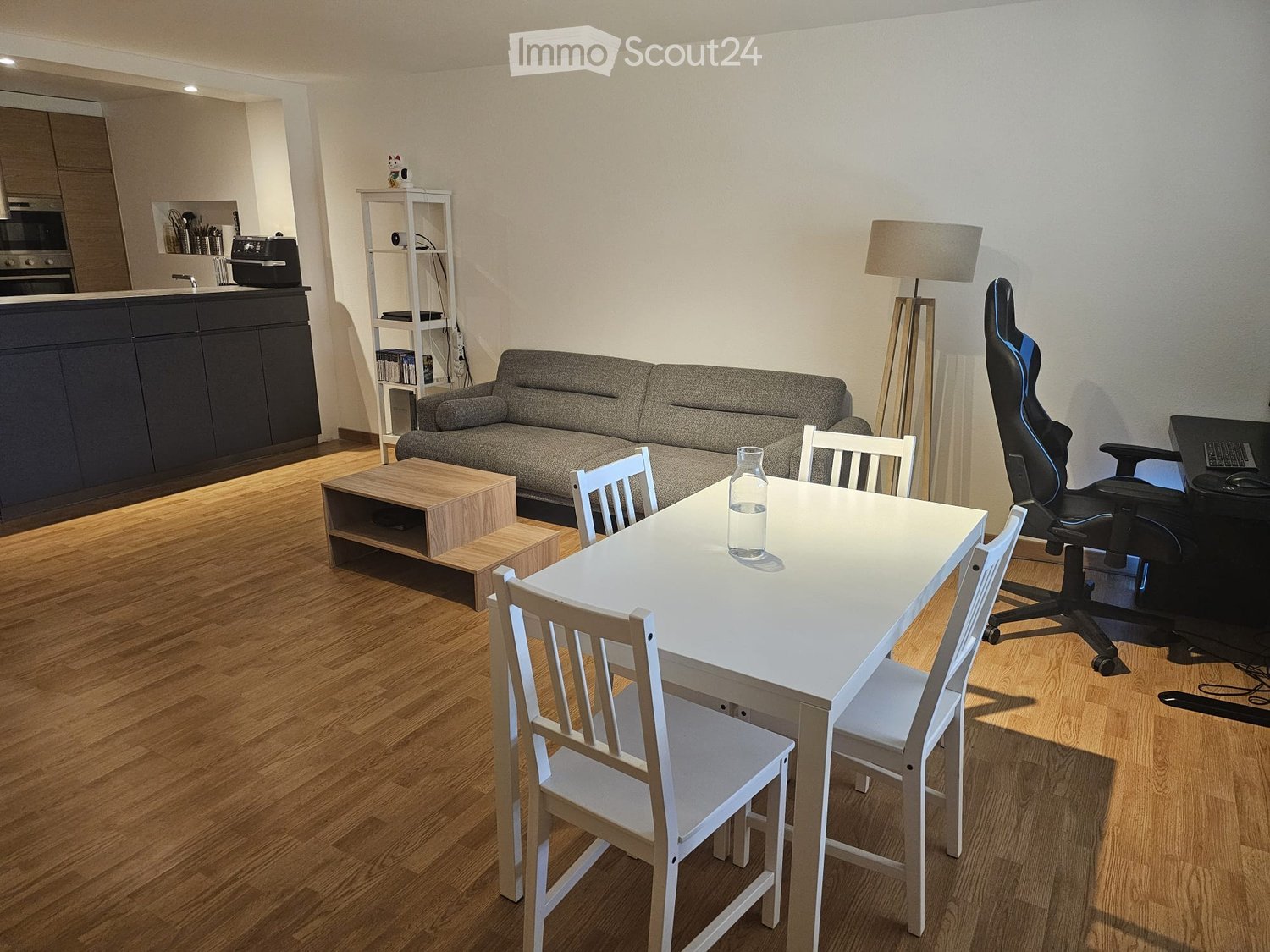 Modern living and dining area with a grey sofa, wooden coffee table, white dining table with chairs, floor lamp, and a small shelf with items. The space features a kitchen area with dark cabinets and appliances.