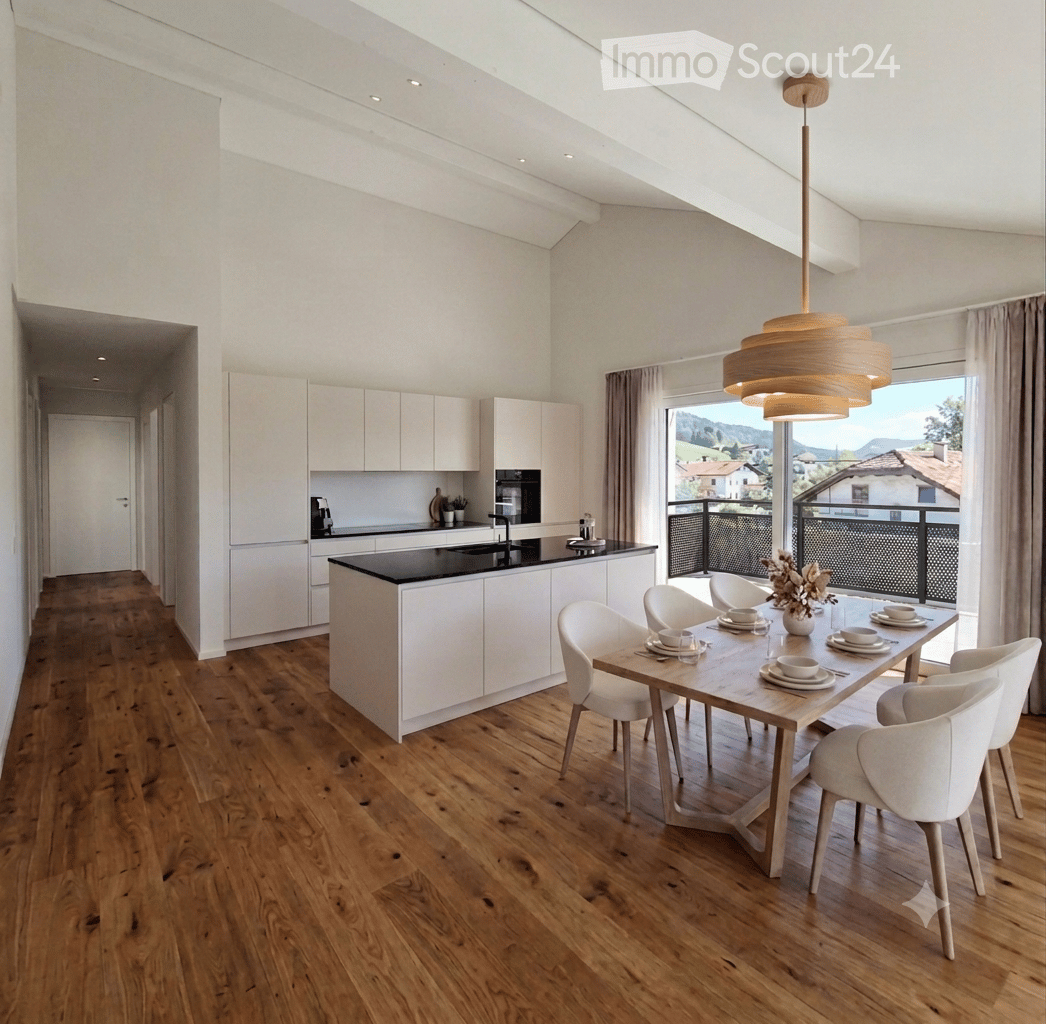 Open-plan kitchen and dining area with light wood flooring, white cabinets, a central island, and a dining table with six chairs. A pendant light hangs above the table, and a large window offers a scenic view.