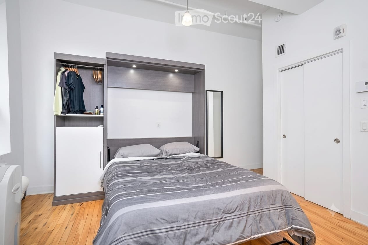 Modern bedroom with a grey duvet-covered bed, built-in wardrobe with hanging clothes, a small refrigerator, and a mirror on the wall.