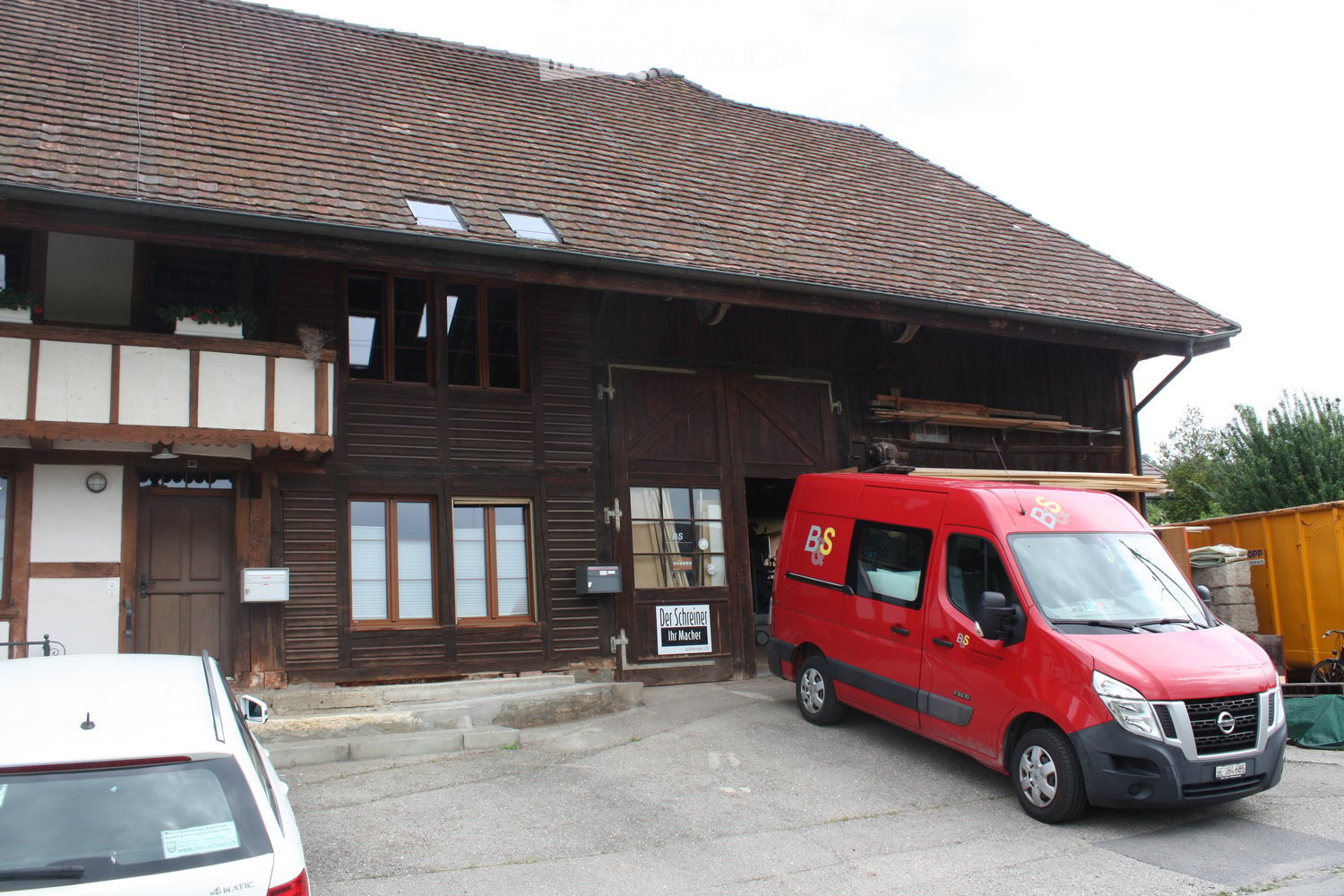 Traditional wooden building with a steeply pitched roof, large windows, and a balcony. A red van is parked in front, and there is a sign on the building.