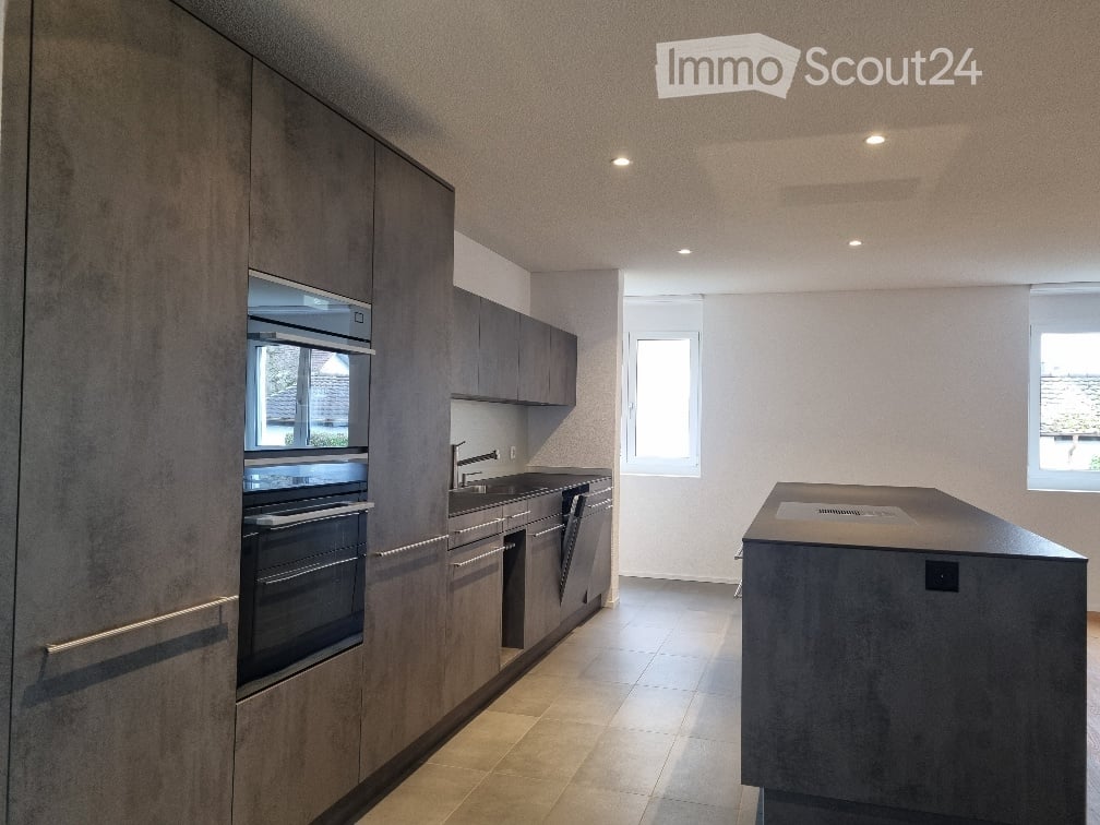 Modern kitchen with dark grey cabinets, built-in oven, sink, and a central island. The space features recessed lighting and large windows allowing natural light to fill the room.
