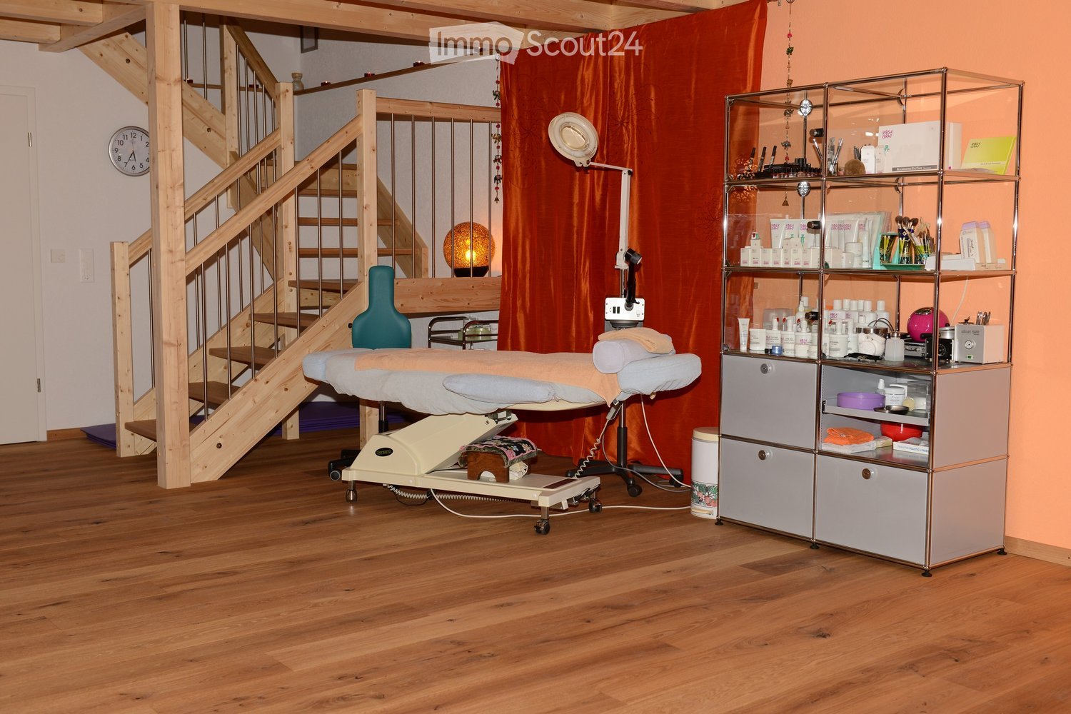 A well-lit, wooden-floored room with a massage table, a metal cabinet filled with various items, a small stool, and a decorative lamp. The room features a staircase leading to an upper level and a large orange curtain in the background.
