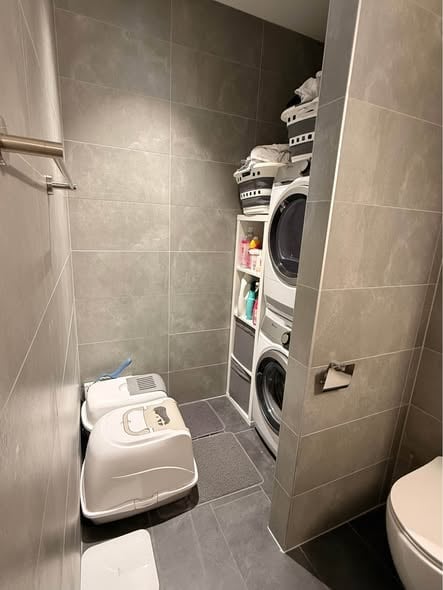 Modern bathroom with grey tiled walls and floor, featuring a toilet, washing machine, laundry basket, and cat litter box.