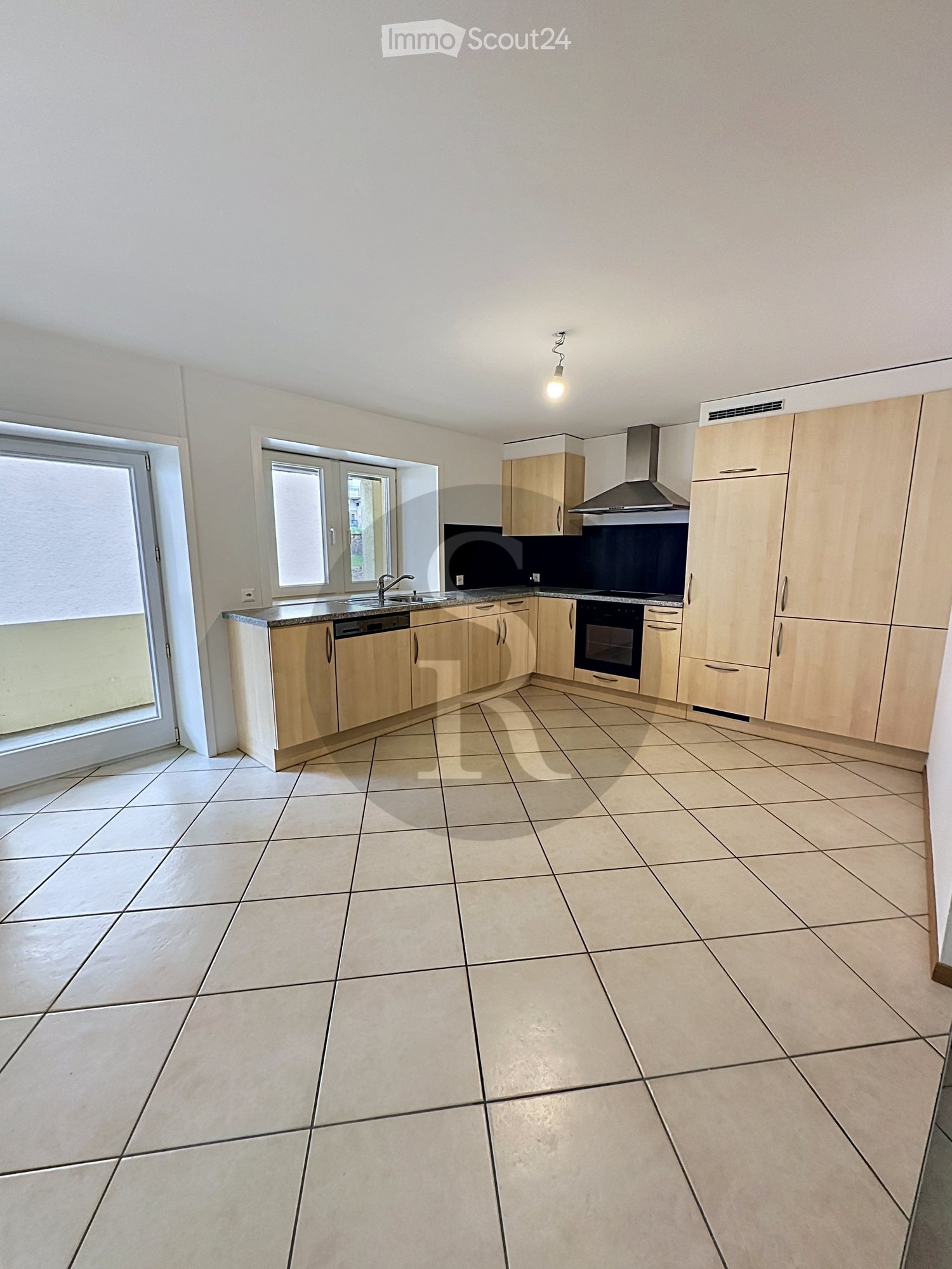 Empty kitchen with light wooden cabinets, tiled floor, and built-in appliances including a sink, dishwasher, and oven. Windows provide natural light.