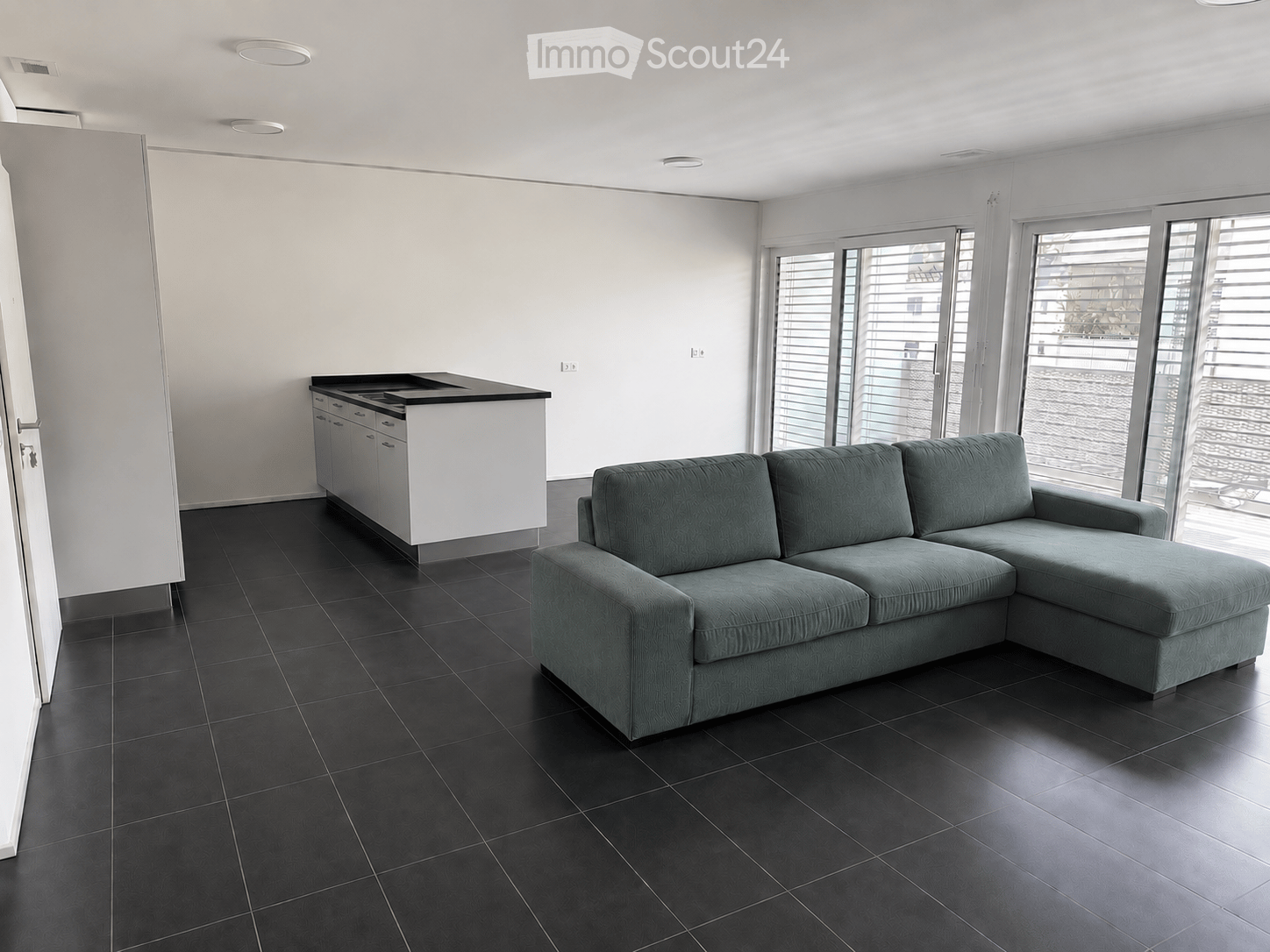 Modern living room with a grey sectional sofa, black tiled floor, white kitchen island with cabinets, and large windows with blinds.