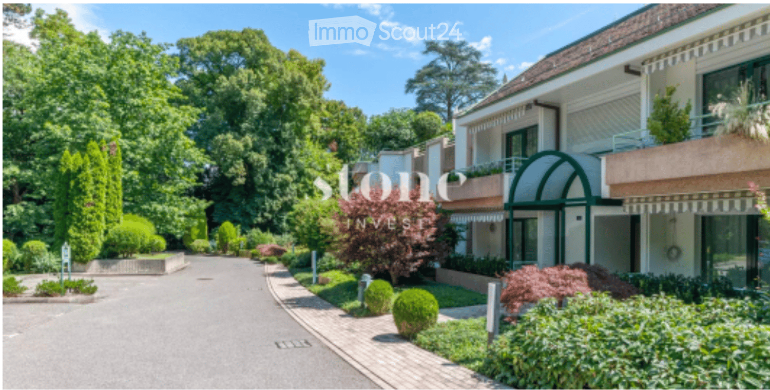 A modern multi-story building with a well-maintained garden and driveway, featuring balconies and large windows.