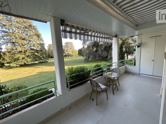 Balcony with white tiled floor, green metal railing, striped awning, small round glass table, and two wicker chairs. Offers a view of a green lawn and trees.