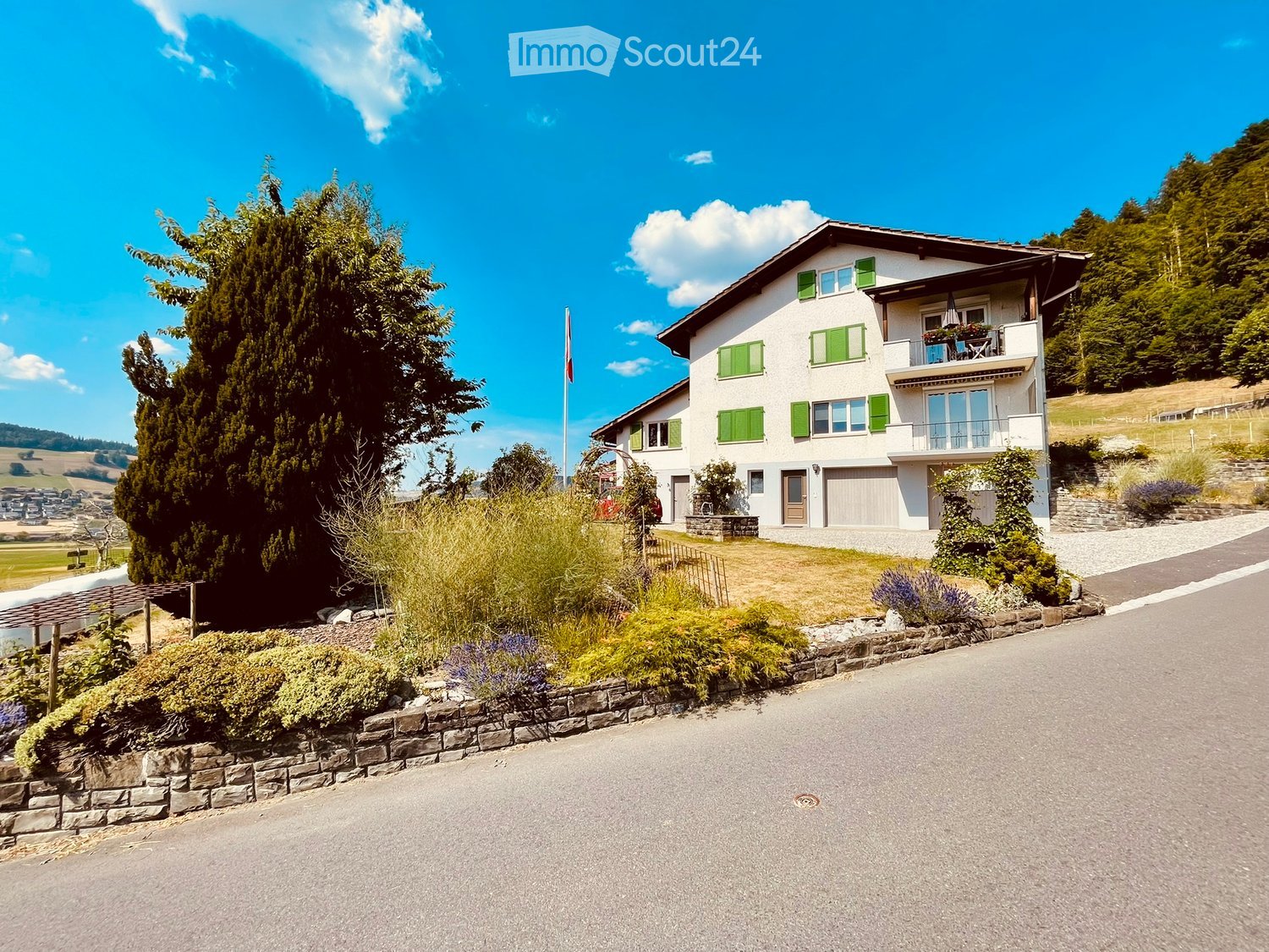 Two-story white house with green shutters, garage, and balcony overlooking a scenic countryside with trees and a clear blue sky.