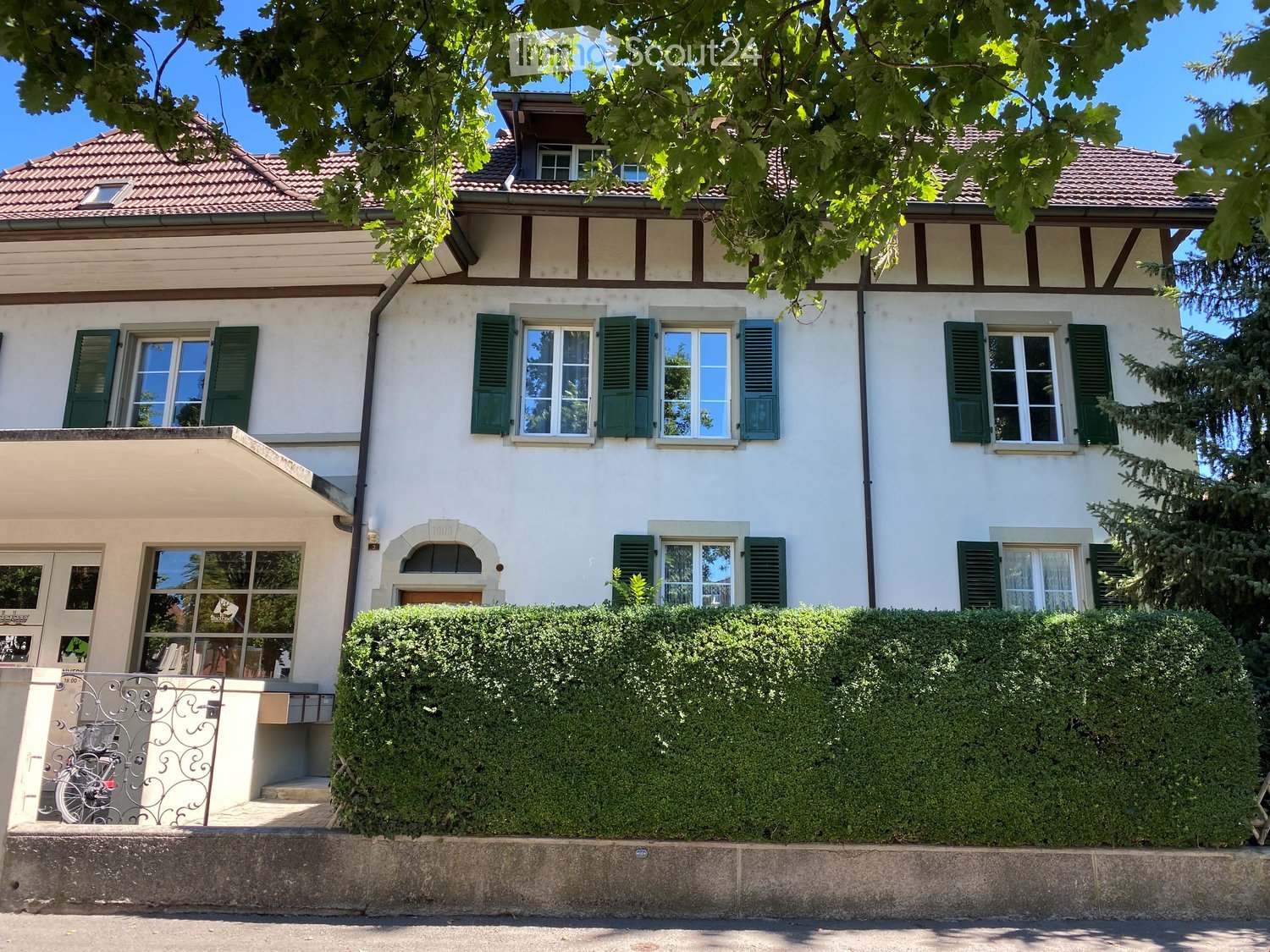 Two-story house with white walls, green shutters, and a red-tiled roof. Features a well-maintained hedge in front and a bicycle parked near the entrance.