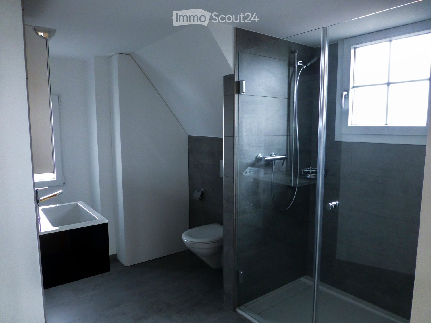 Modern bathroom with a glass-enclosed shower, a white sink with a black cabinet, and a toilet. The room features a slanted ceiling and large windows allowing natural light.