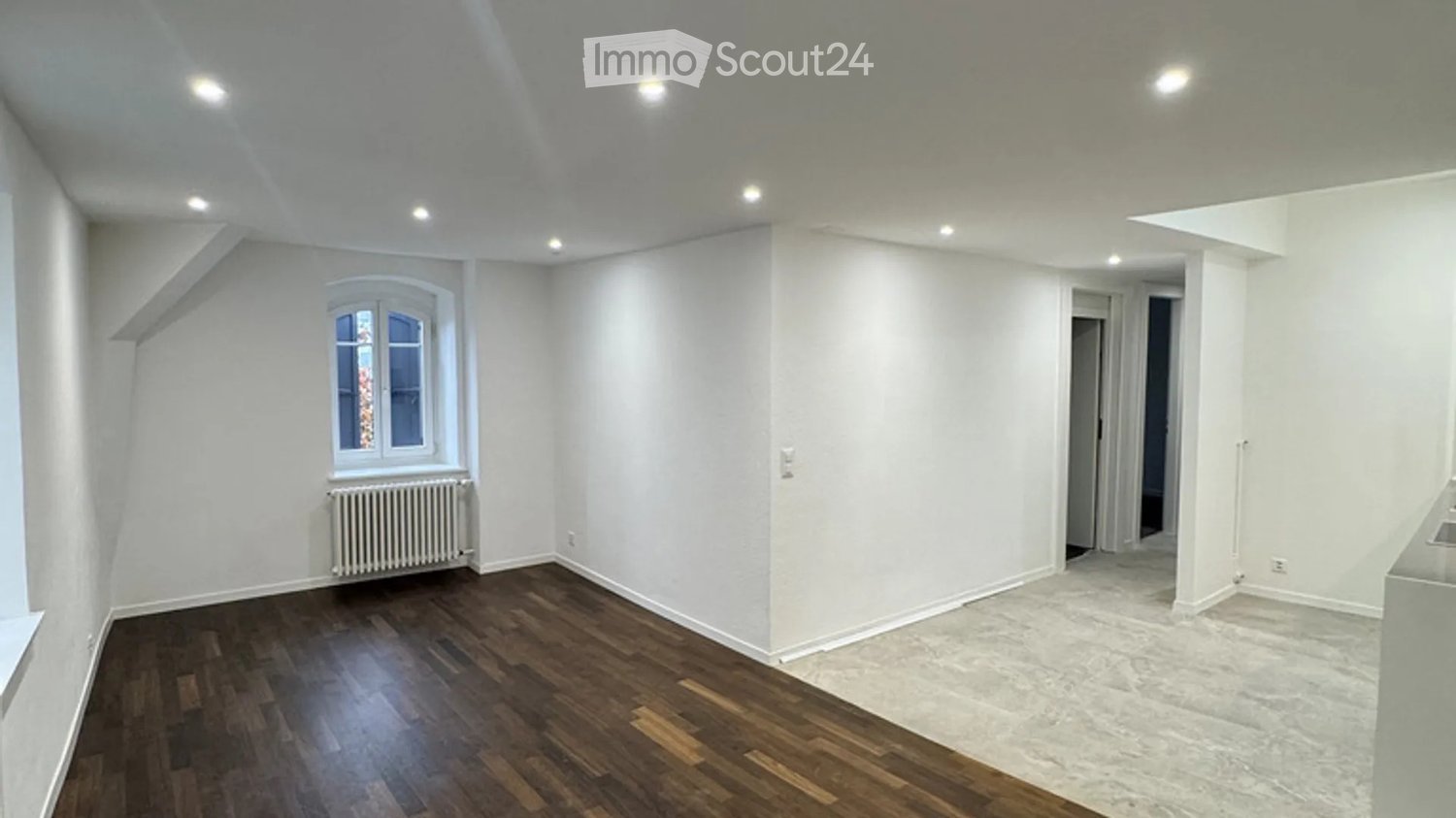 Empty room with wooden flooring, white walls, and a window with a radiator below it. The room has recessed lighting and an open doorway leading to another space.