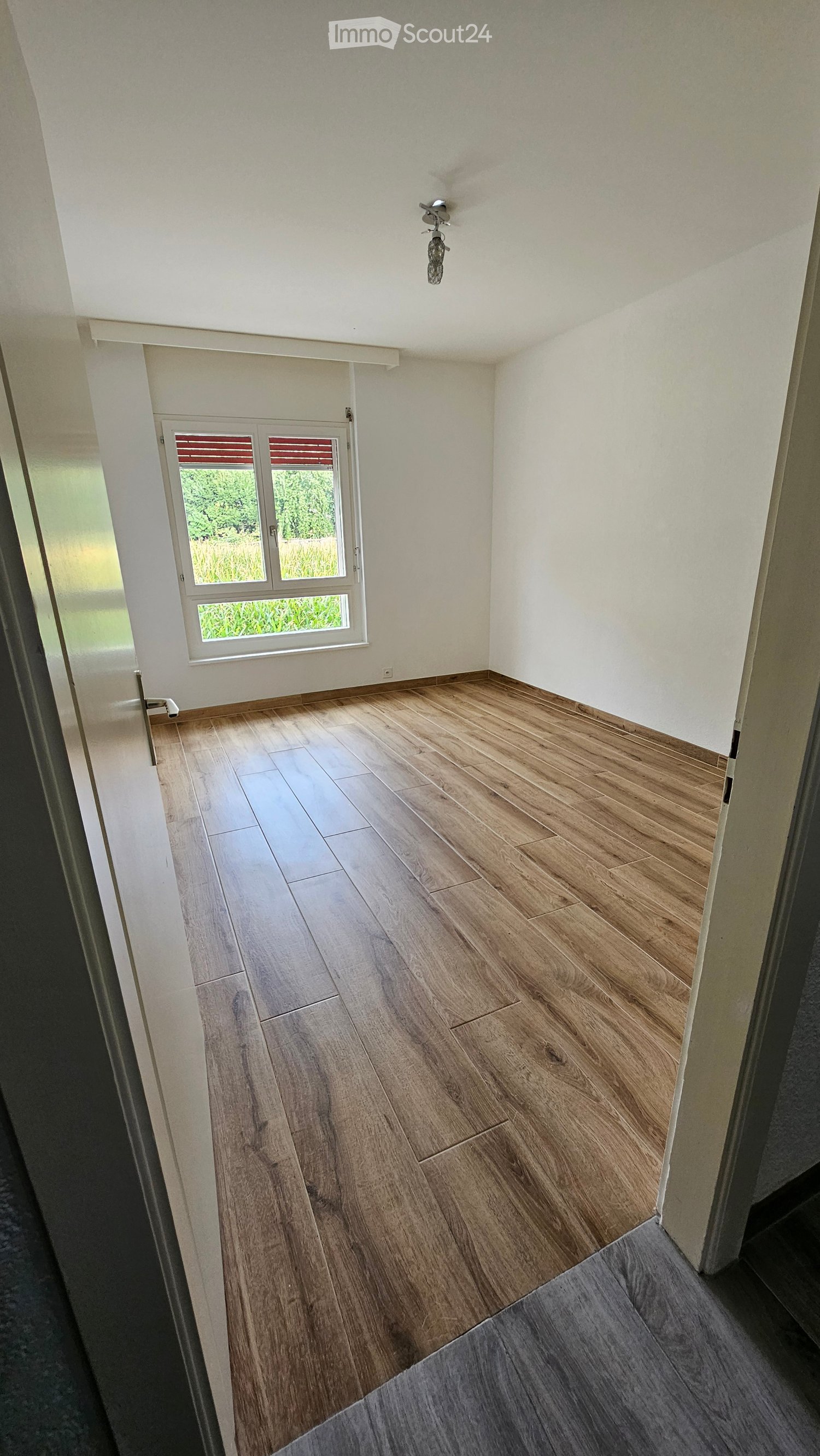 Empty room with wooden flooring and white walls, featuring a window with blinds and a door leading to another space.