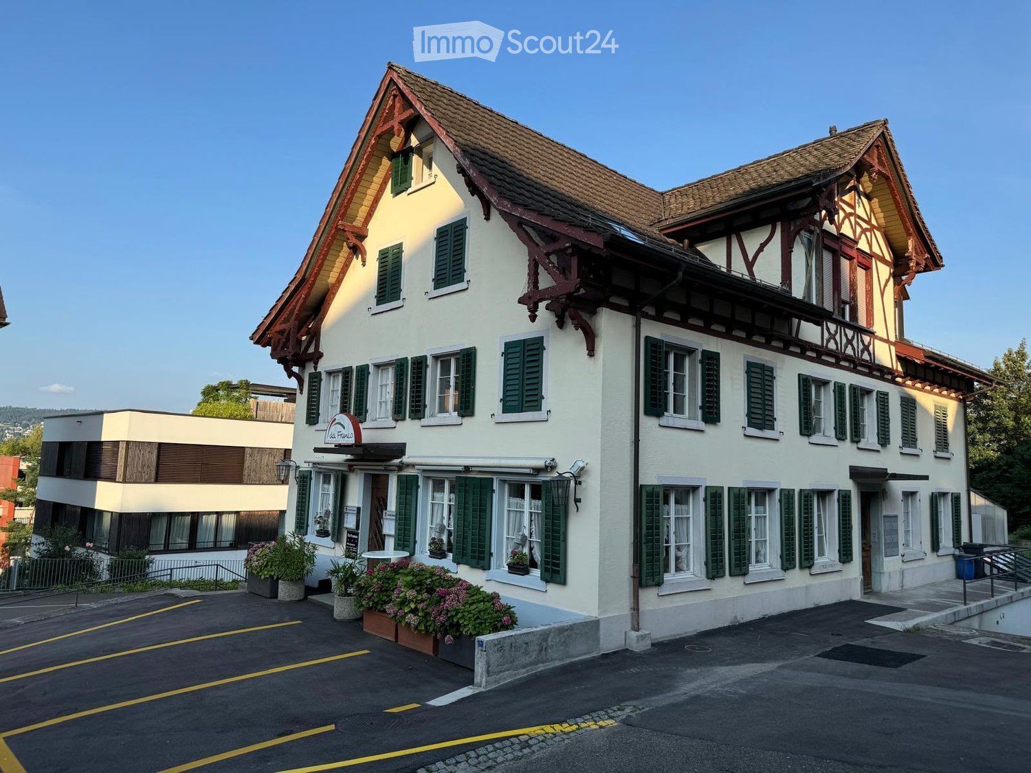 Two-story traditional building with white walls, green shutters, and a tiled roof. The ground floor features a restaurant sign and flower boxes, with a modern adjacent building and parking area.