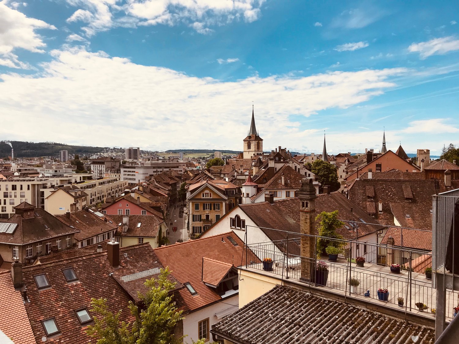 Balcony overlooking a cityscape with traditional rooftops, chimneys, and distant spires under a partly cloudy sky.