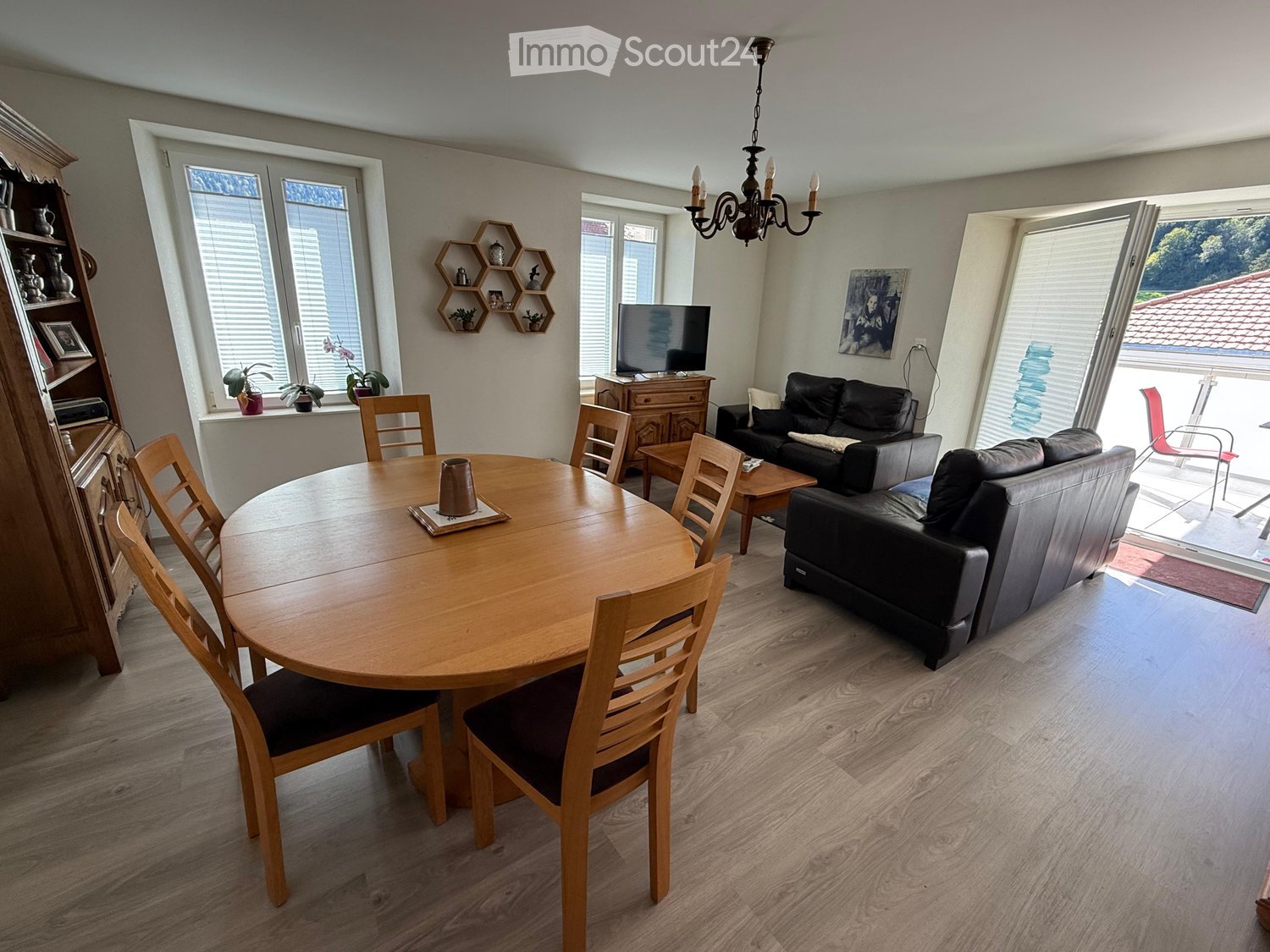 Bright living and dining area with wooden flooring, a round wooden dining table with chairs, a black leather sofa, a TV on a cabinet, and a chandelier hanging from the ceiling. Large windows and a sliding glass door lead to a balcony.