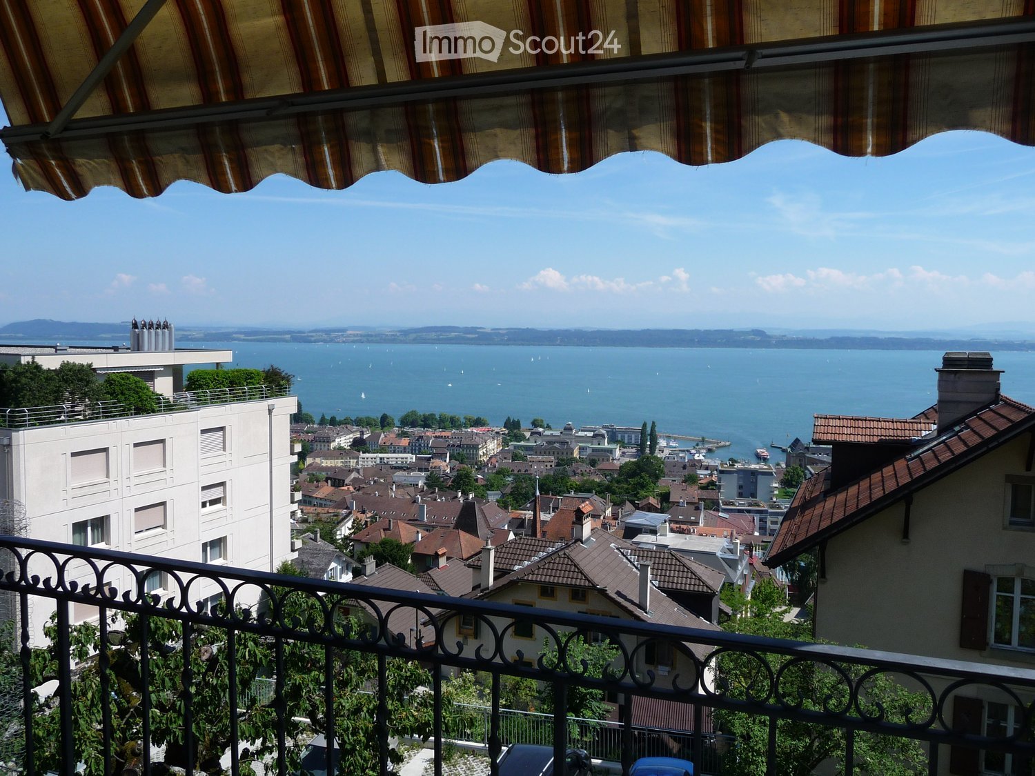 Balcony with metal railing, striped awning, overlooking a cityscape with rooftops, a lake, and distant mountains.