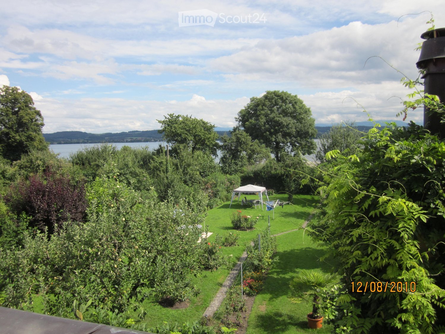 A well-maintained garden with a lawn, trees, a small white tent, and a view of a lake and distant mountains.